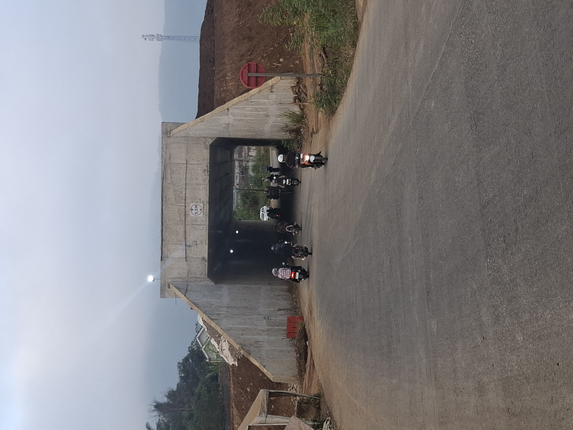 This image depicts a road leading towards an underpass in Ciheulang Tonggoh, Indonesia. The underpass is constructed of concrete with a sign indicating a height limit of 5.5 meters. Several motorcycles, with riders, are entering and passing through the underpass. The surrounding environment features piles of earth and construction materials, suggesting ongoing or recent infrastructure work. Vegetation is visible on the slopes and to the sides of the road, and in the background, a telecommunications tower stands against a hazy sky. The overall atmosphere is overcast, characteristic of the Indonesian climate.