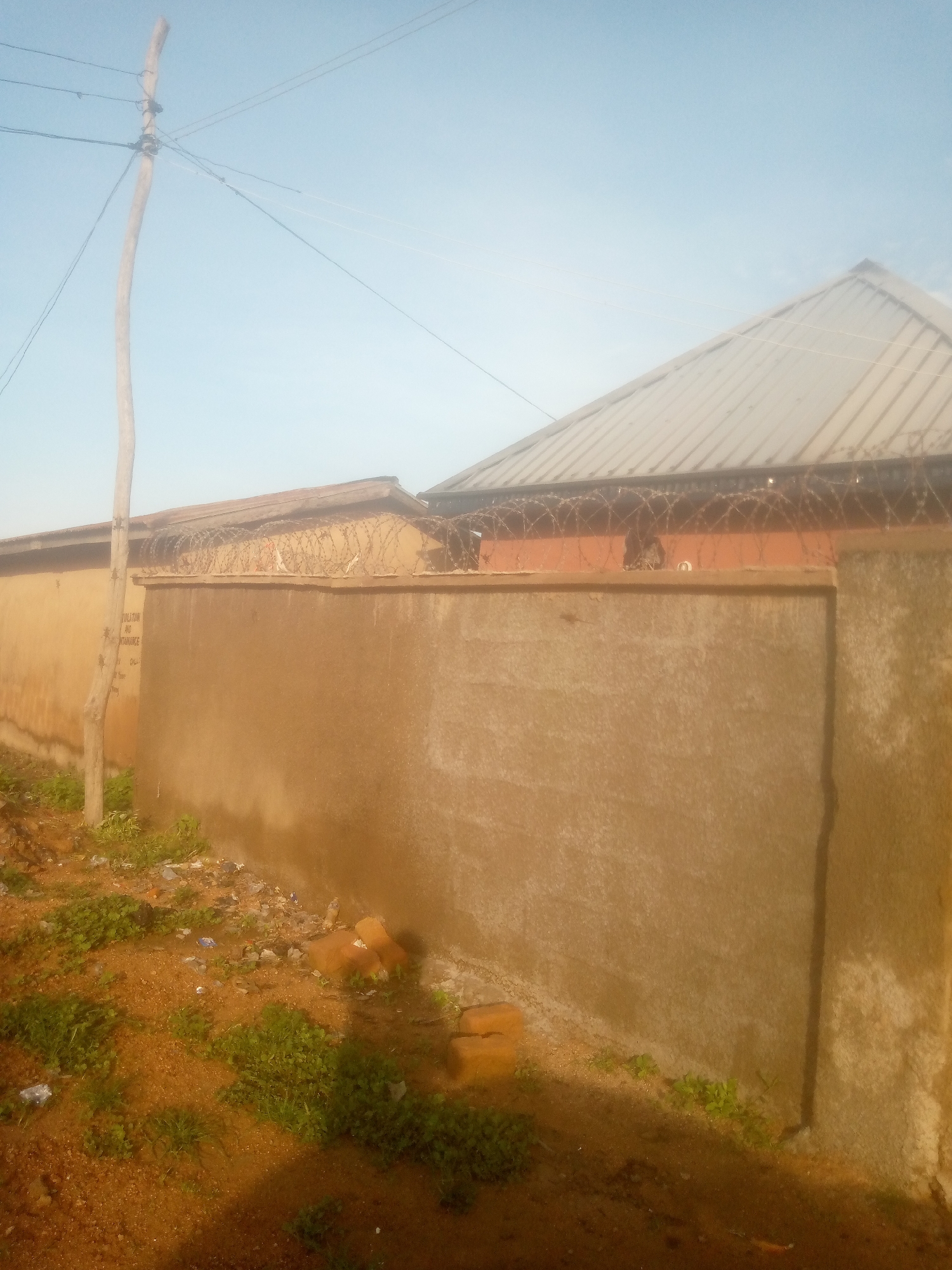 The image depicts a property in Jalingo, Nigeria, enclosed by a tall, rough-textured concrete wall topped with barbed wire. A portion of a corrugated metal roof of a building is visible over the wall. To the left, a wooden utility pole with electrical wires is prominent. The ground in front of the wall is a mix of dirt and sparse vegetation, with some scattered debris. The overall scene suggests a residential or commercial property with security measures in place.