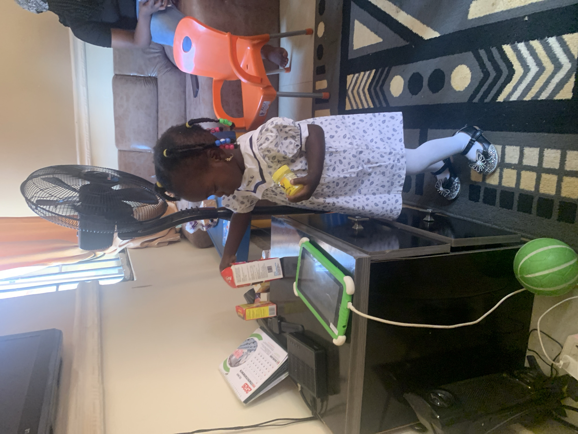 In a room in Gura Top, Nigeria, a young girl stands near a table with a tablet. The girl, wearing a white dress with a blue trim and a white bow, is holding a yellow bottle. On the table, a tablet with a green case is plugged in. To the left of the tablet, a black keyboard and a calendar are visible. A large black fan is positioned behind the girl. In the background, a brown couch and an orange chair are present. A woman is seated on the couch, wearing blue jeans and a black shirt. The room is decorated with a patterned rug and has windows with orange curtains.