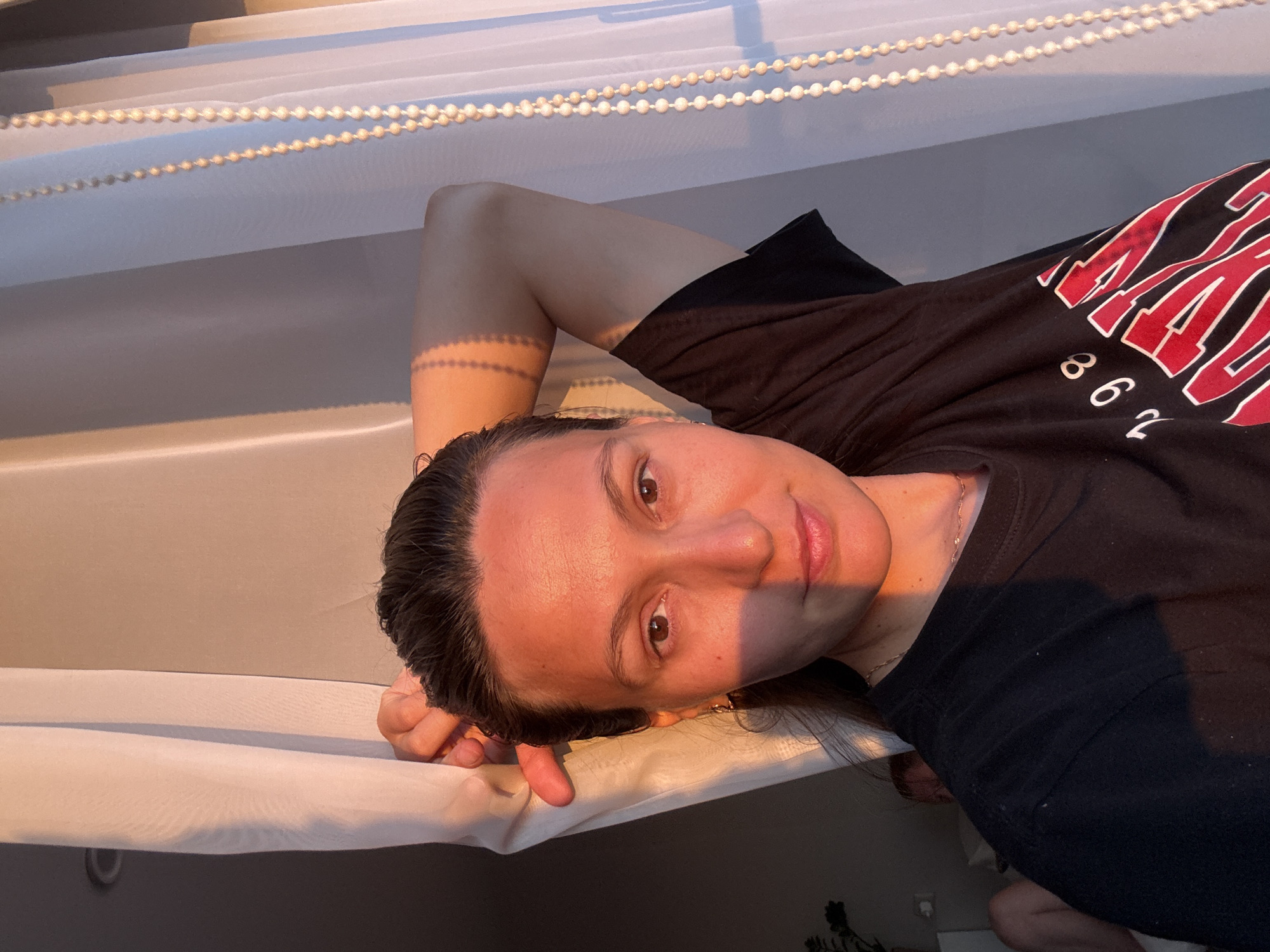 The image shows a woman with her arm raised behind her head, looking towards the camera. She is wearing a black t-shirt with red and white lettering. Her face is partially illuminated by warm, golden light, suggesting it is either sunrise or sunset. To her right, a sheer white curtain with beaded trim hangs near a window. The background is dimly lit and appears to be an indoor setting. A blurred figure can be seen in the background to the left, suggesting another person is present. The scene does not contain any identifiable landmarks or features that would place it in Chaikovsky, Russia.