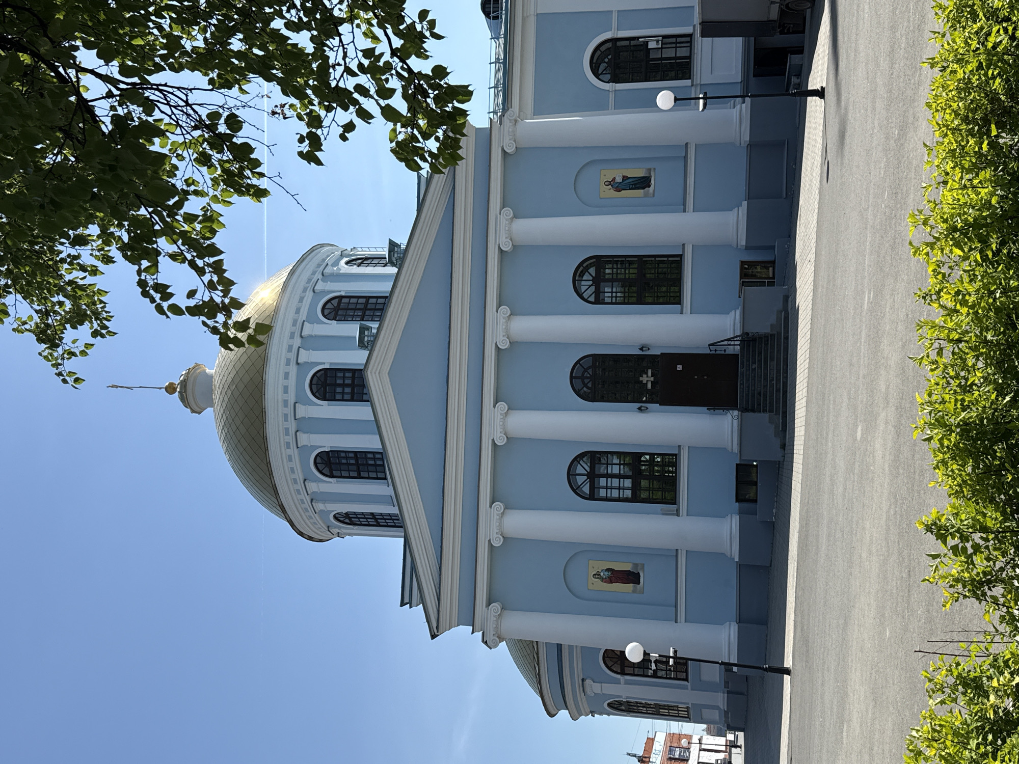 The image captures a section of the Cathedral of the Annunciation in Votkinsk, Russia. The lower portion of the building is pale blue with white columns. There are rectangular windows on the building's sides and portraits between the columns. A golden dome is visible, along with part of the roof. Part of the road is visible, and the image's right and left edges contain tree foliage. The sky is pale blue.