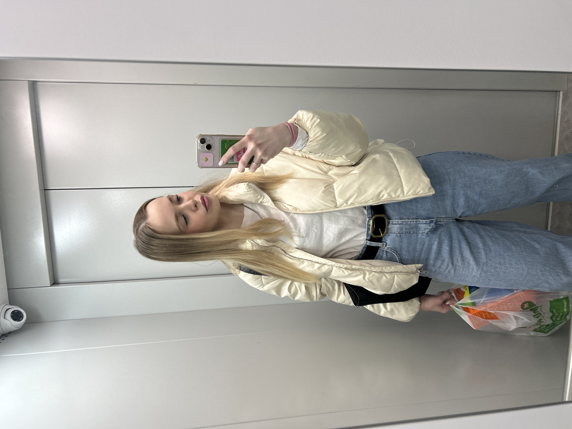 A fair-skinned, blond woman is taking a selfie in a reflective elevator. She wears a white puffy jacket, white t-shirt, blue jeans with a black belt, and a pink bracelet. She holds a plastic shopping bag in her left hand and a smartphone in her right. The elevator interior consists of gray walls, metal flooring, and a security camera in the upper left corner. This image was taken in the city of Chaikovsky, Russia.