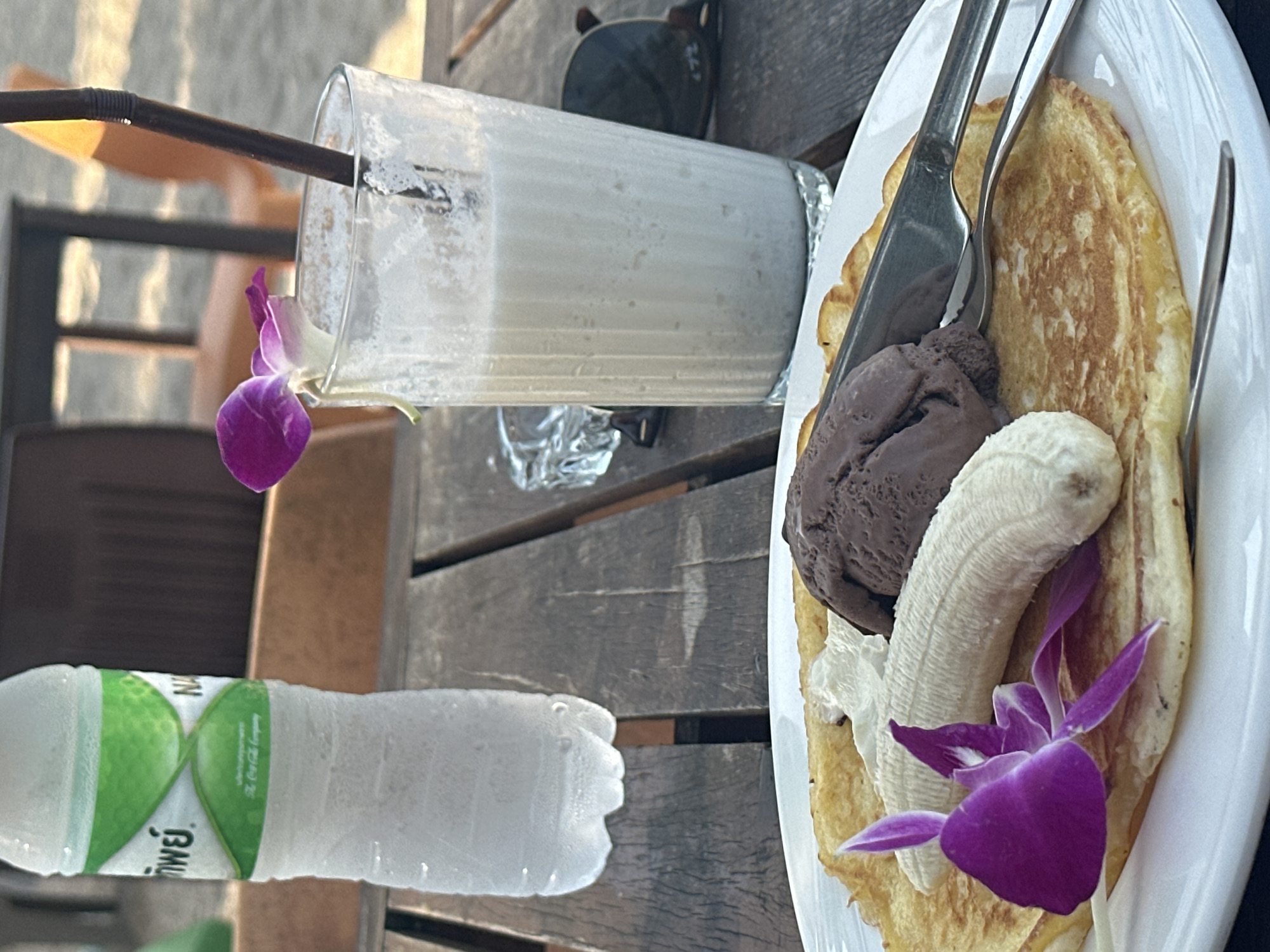 The scene captures a still life of refreshment and indulgence on a wooden table, potentially at a beachside cafe in Baan Chaweng Noi, Thailand. A partially consumed creamy smoothie stands in a tall glass with a straw. Adjacent to it, a plate of pancakes topped with a scoop of chocolate ice cream and a banana slice, garnished with a vibrant purple orchid, suggests a sweet treat. Silverware is laid out alongside the plate. Nearby, a bottle of water bears a green label with Thai script. Sunglasses rest casually on the table, hinting at a sunny day. The setting implies a relaxed vacation atmosphere.