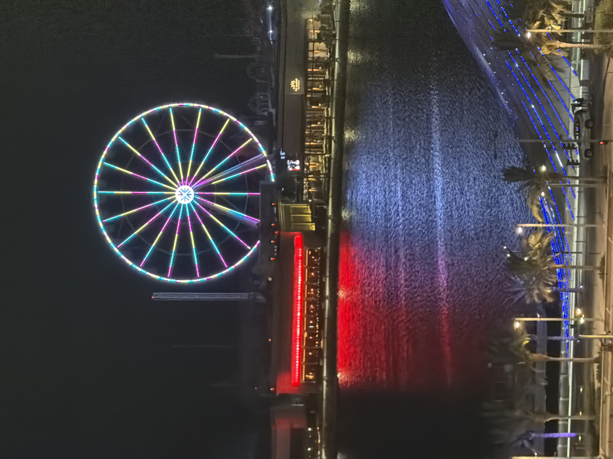 This nighttime image depicts an illuminated entertainment complex alongside a body of water, observed from an elevated perspective. A prominent, large Ferris wheel dominates the upper center, radiating multi-colored lights in a radial pattern (teal, yellow, pink, purple) and along its outer rim. To the left of the Ferris wheel, a building features a striking horizontal red light array that casts a strong, vibrant reflection onto the water. To the right, another building with warm, inviting interior lighting and a sign displaying "THE BEACH" (partially legible) also reflects in the water. The water surface, likely a lake or canal, exhibits ripples and accurately reflects the intense red and more subdued warm lights from the buildings, and a faint reflection of the Ferris wheel's colors. In the lower foreground, a paved area or road runs parallel to the water's edge, lined with several palm trees. Multiple streetlights illuminate this area. A light-colored vehicle, possibly an SUV, is parked on the road, with at least four indistinct human figures standing near it on the right side. A blue-lit architectural feature, possibly a ramp or wall, is visible on the lower right, casting blue light onto the ground and water. The sky is dark and clear. The location in the city is unknown.