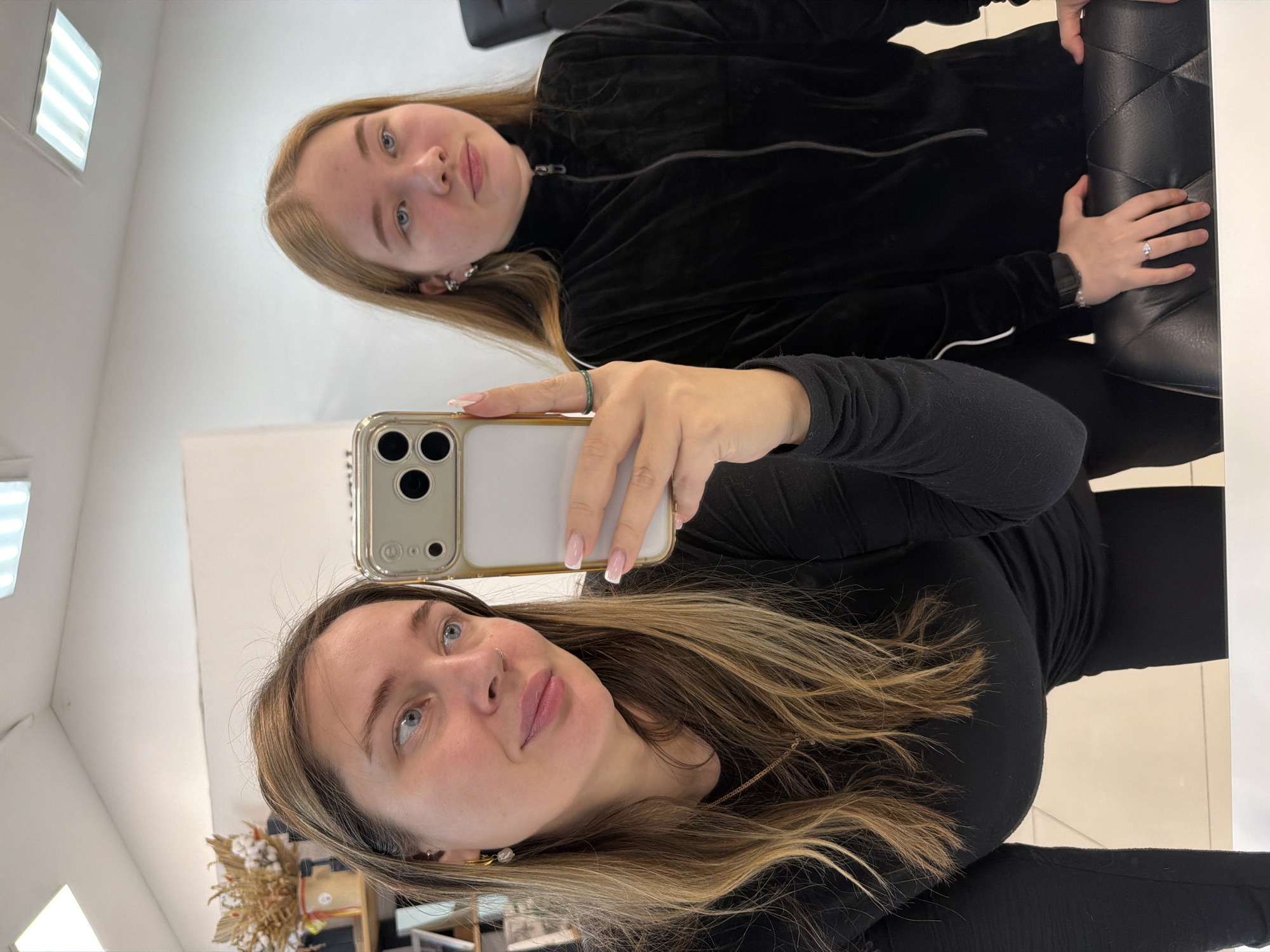The image shows two females posing for a mirror selfie inside a well-lit room in Chaikovsky, Russia. The individual on the left, an adult female with long, light brown hair with blonde highlights, blue eyes, and fair skin, is holding a light-colored smartphone with a multi-lens camera in her left hand, pointed towards the mirror, indicating she is taking the photograph. She wears a black long-sleeved top, gold hoop earrings with white pearl-like drops, a thin gold necklace, and a small nose ring. Her right hand, displaying light pink French-tip nail polish, features a green ring on the index finger. She has a subtle smile and directs her gaze towards the phone's screen.
Behind and to the right stands a younger female with long, light brown hair and blue eyes. She is wearing a black velour zip-up jacket with a white stripe visible on the inner arm. Her left ear is adorned with multiple small earrings, and a dark smartwatch is visible on her left wrist. Her right hand rests on the back of a black, quilted upholstered chair. She maintains a neutral expression, looking directly at the camera/mirror. The background features white walls and ceiling, overhead square fluorescent light fixtures, and on the left, a partial view of a shelf unit displaying a decorative arrangement of dried plants.