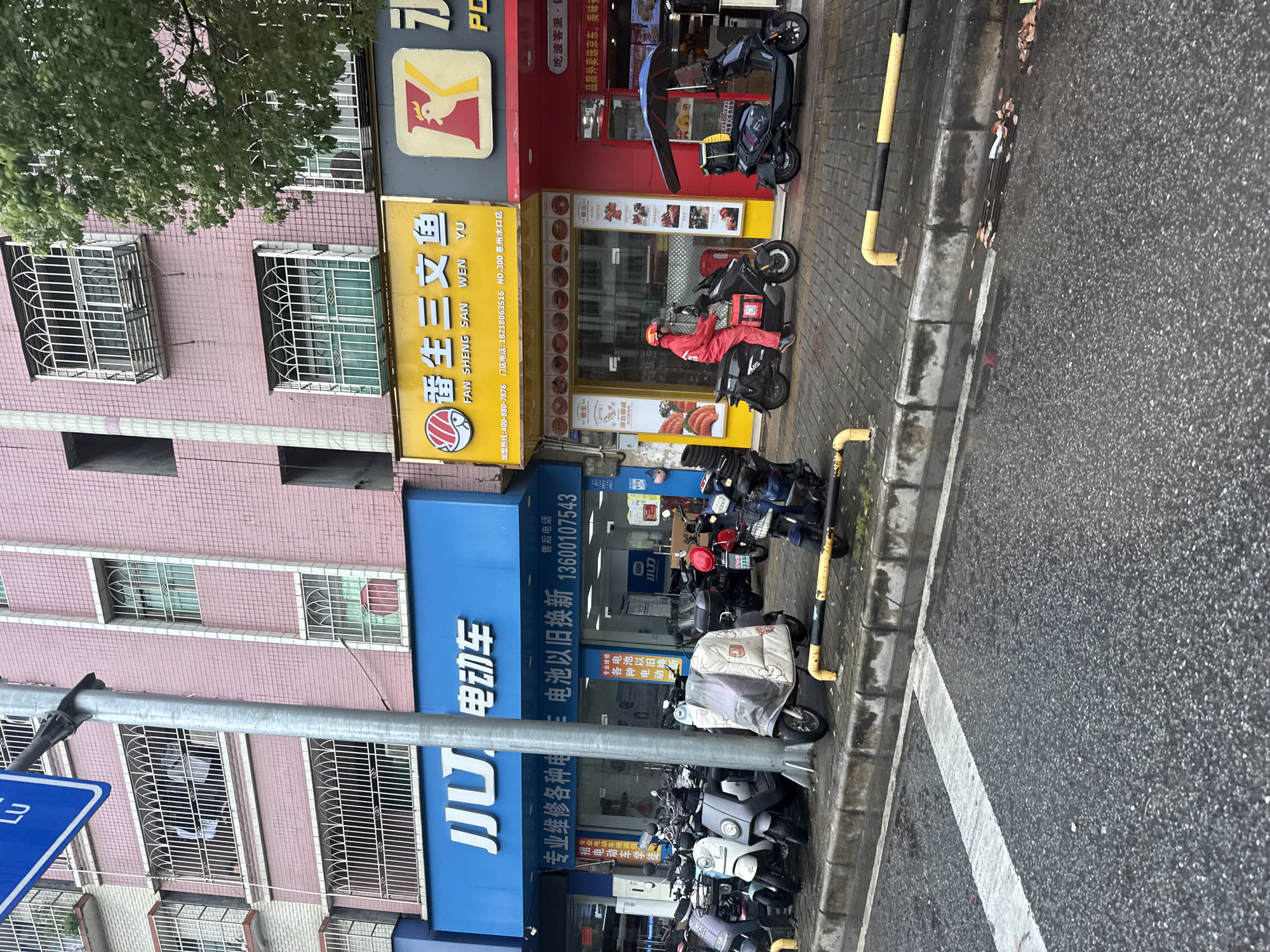 This is a street-level, eye-level photograph of a commercial street in Huizhou, China. The scene depicts a row of storefronts, primarily housing businesses related to electric vehicles and food. In the foreground, a wet asphalt road with a white line marking a lane is visible. A concrete curb separates the road from the sidewalk, which is paved with interlocking bricks and lined with metal barriers.
Along the sidewalk, a collection of parked electric scooters and motorcycles are visible, some covered with protective cloths. One scooter in the middle foreground is partially covered with a patterned cloth.
The buildings have a facade of pink tiles, with several windows on upper floors fitted with white metal grilles. Several businesses are identifiable by their signage. A prominent blue sign reads "JJ.U 电动车," indicating an electric vehicle store, which also advertises services like "professional repair of various electric vehicles" and "battery trade-in/replacement." Next to it, a yellow sign with Chinese characters and "FAN SHENG SAN WEN YU" written in English, suggests a food establishment. To the right, a red storefront with a yellow "K" logo, possibly another food business, is visible.
A person wearing a red uniform and helmet, likely a delivery driver, is seated on a black electric scooter in front of one of the food establishments. Another dark-colored electric scooter with a blue umbrella is parked nearby. The overall environment appears to be an urban commercial area, possibly in the morning or after rain, given the wet surfaces.