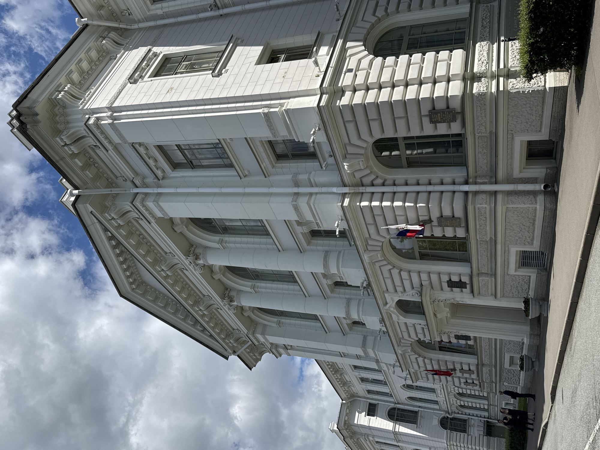 This image shows a tall, ornate white building with classical architectural details, including columns, arched windows, and decorative moldings. The building appears to be a public or institutional structure, possibly a government building or museum. Two flags, one with red, white, and blue stripes and another with the same colors arranged in a different pattern, are visible flying from the building. There are also several people walking on the sidewalk in front of the building. The sky is partly cloudy, suggesting it is daytime. The building's facade is detailed with many windows, some of which appear to be arched, and stone carvings. There are also planters with greenery outside the building. The image is taken from a low angle, looking up at the building. The context of Sinyavino, Russia, suggests this is likely a historical or culturally significant building within that region.