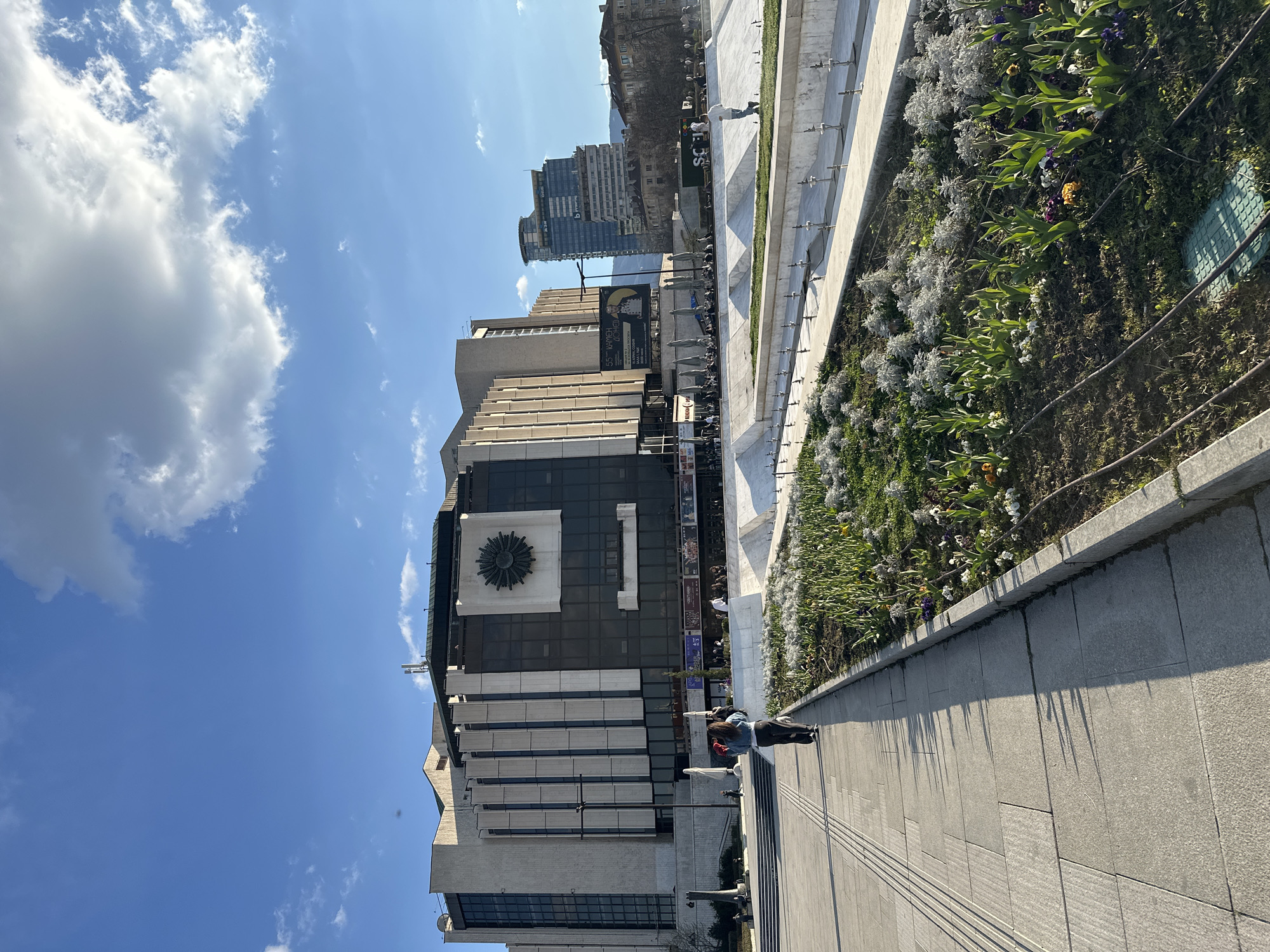 The image depicts a street scene in Sofia, Bulgaria, featuring the National Palace of Culture (NDK) on a sunny day. The NDK building is a large, modern structure with a clock face on its facade. In front of the building, there's a wide pedestrian walkway paved with gray stone, along which people are walking and sitting. A raised flowerbed with greenery and flowers runs parallel to the walkway. In the background, other buildings are visible, including a taller, modern building. The sky is blue with some white clouds. The image appears to be taken from a slightly low angle.