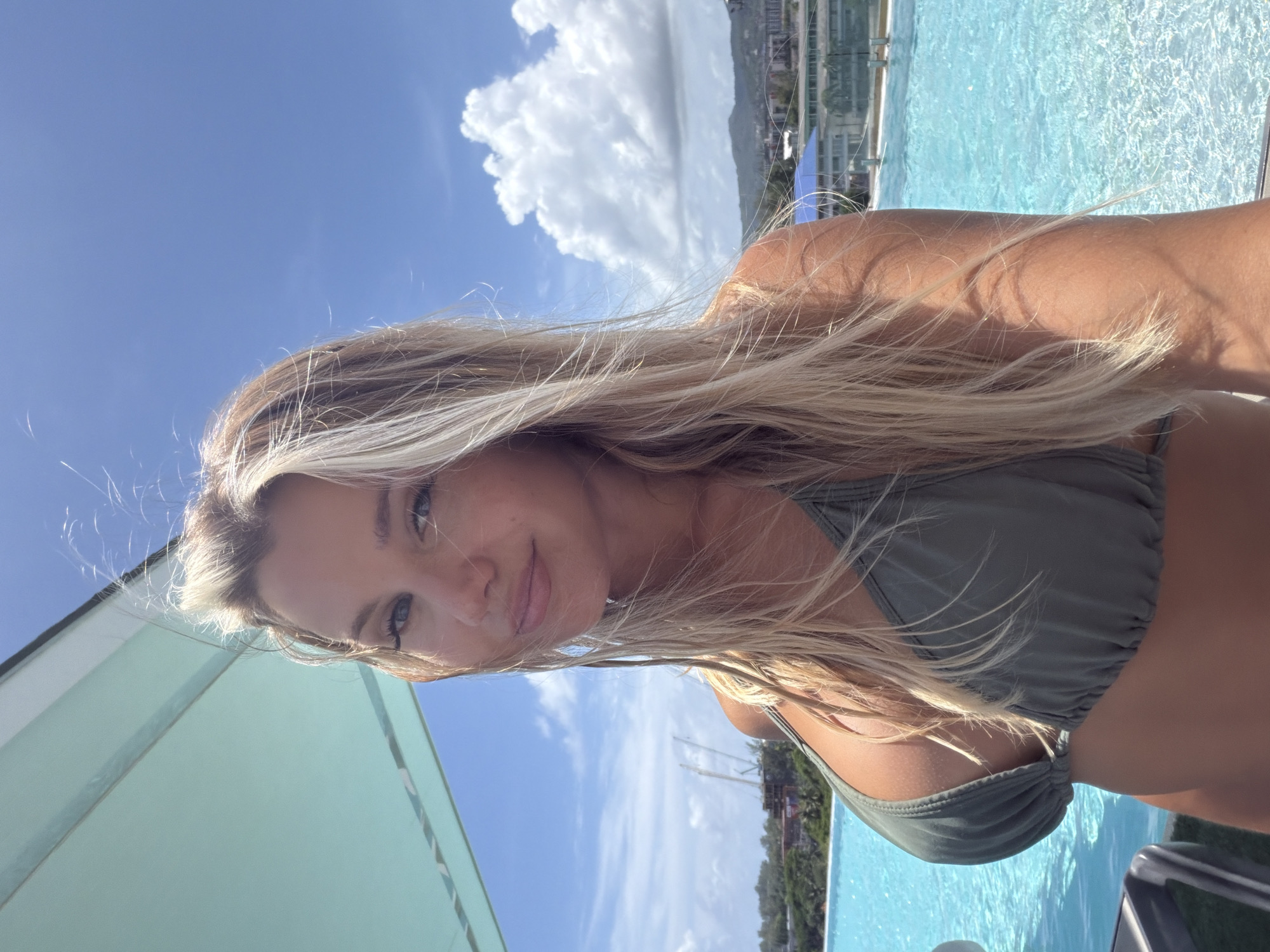 An outdoor, eye-level, medium shot shows a blonde woman in a green bikini top posing for a selfie by a swimming pool. The woman is looking at the camera with her head tilted slightly to her right. She has long, blonde hair that is blowing in the wind. The swimming pool is blue and has clear water. In the background, there are buildings and trees. The sky is blue with some clouds. The image appears to have been taken on a sunny day.