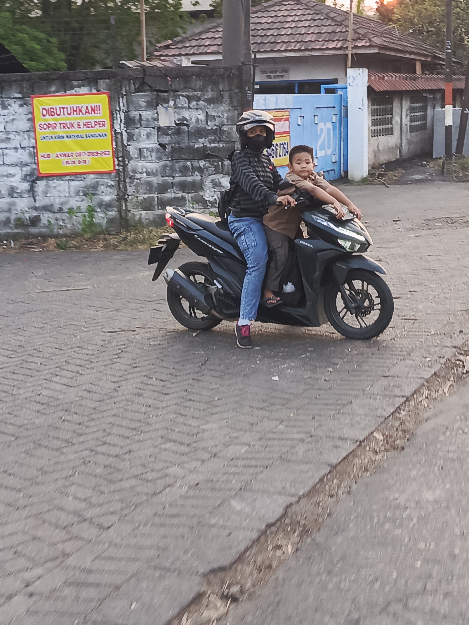 A woman and a young boy are riding a black motorcycle on a paved road. The woman is driving and is wearing a helmet and a black face mask. She has on a striped jacket and blue jeans. The boy is sitting behind her, wearing a brown shirt. They are both looking forward.
The scene appears to be in a residential or industrial area, with a brick wall in the background. A yellow sign with red lettering is attached to the wall. The sign contains text in Indonesian, with the words "DIBUTUHKAN!!" (Needed!!) prominently displayed, followed by "SOPIR TRUK & HELPER" (Truck Driver & Helper). Below this, it states "UNTUK KRIM MATERIAL BANGUNAN" (For Construction Material). Contact information is also listed on the sign.
Behind the wall, a building with a tiled roof and a blue gate with the number "20" is visible. The lighting suggests it is late afternoon or early evening, possibly during sunset, as the sky has a warm glow. The road surface is made of interlocking paving stones. The overall impression is a casual moment of transportation in a developing area.