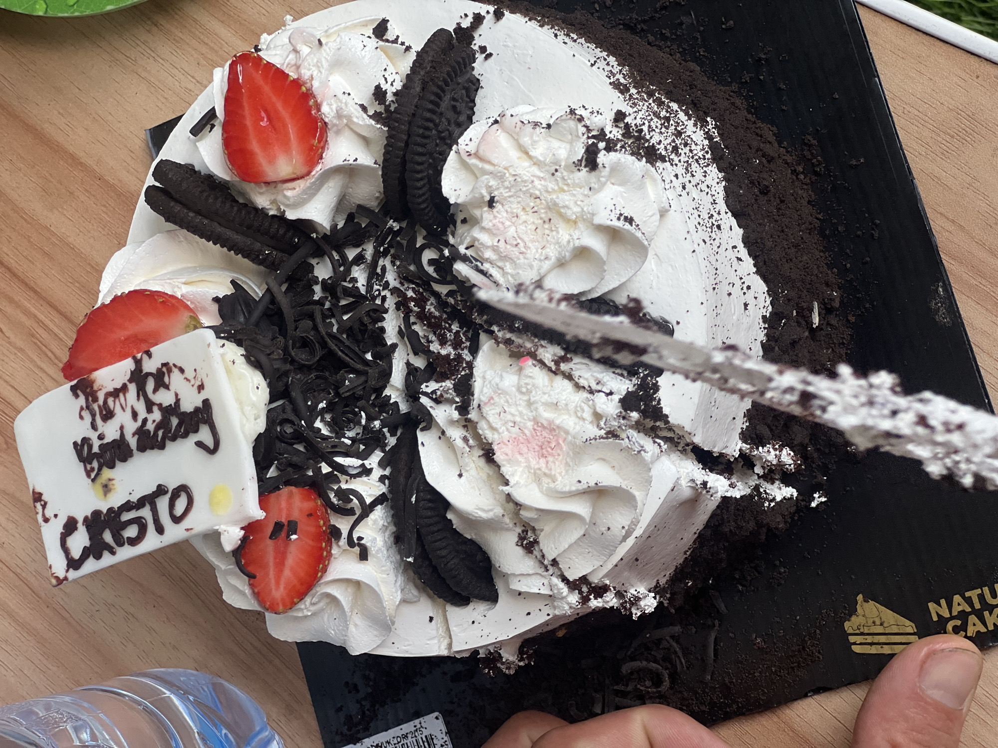 In Denpasar, Indonesia, a slice is being cut from a black forest cake decorated with white frosting, chocolate shavings, strawberries, and cookies. The cake rests on a black platter with a gold logo that reads "NATU CAK." A person's hand is visible, holding a knife as it cuts into the cake. The scene is set on a wooden table, with a bottle of water to the left of the cake. The overall atmosphere suggests a casual celebration or gathering.