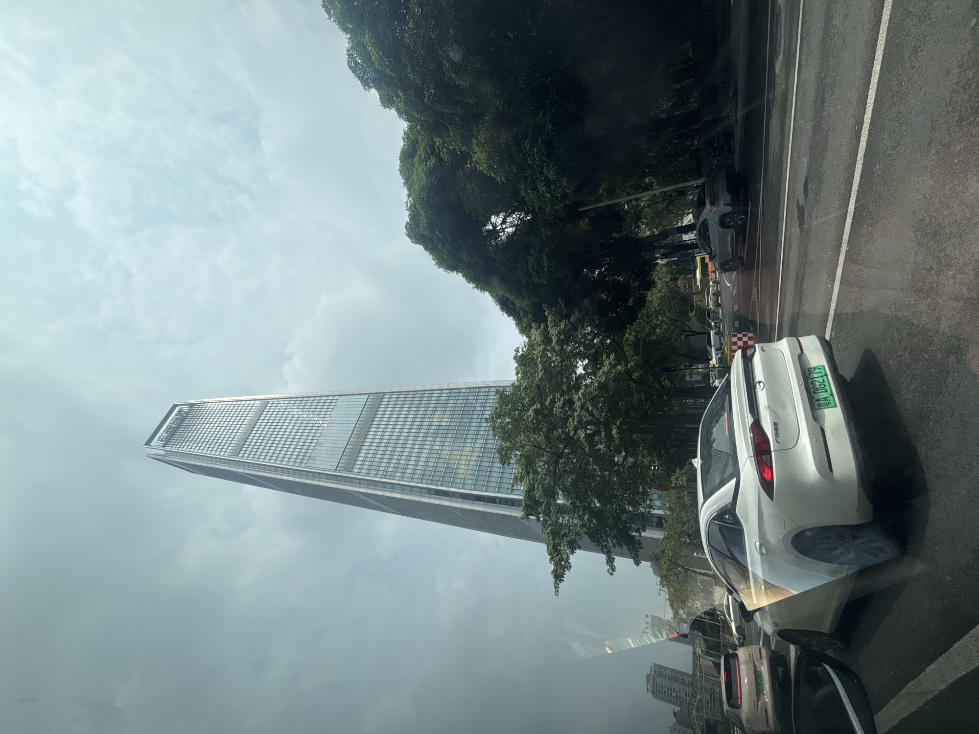 In Tianhe District, China, a tall, modern skyscraper with a glass facade dominates the skyline under a cloudy sky. The skyscraper features a distinctive tapered shape and is adorned with lettering near its peak. In the foreground, traffic moves along a wet asphalt road, indicating recent rain. A white sedan with a green license plate, registered as "粤A DG2179," is prominent in the right lane, facing away from the viewer. Another white car is visible further ahead in the same lane. To the right of the road, a dense canopy of lush green trees lines the street, partially obscuring buildings in the distance. The reflection of traffic and a portion of the skyscraper is visible on the surface of the vehicles in the foreground.