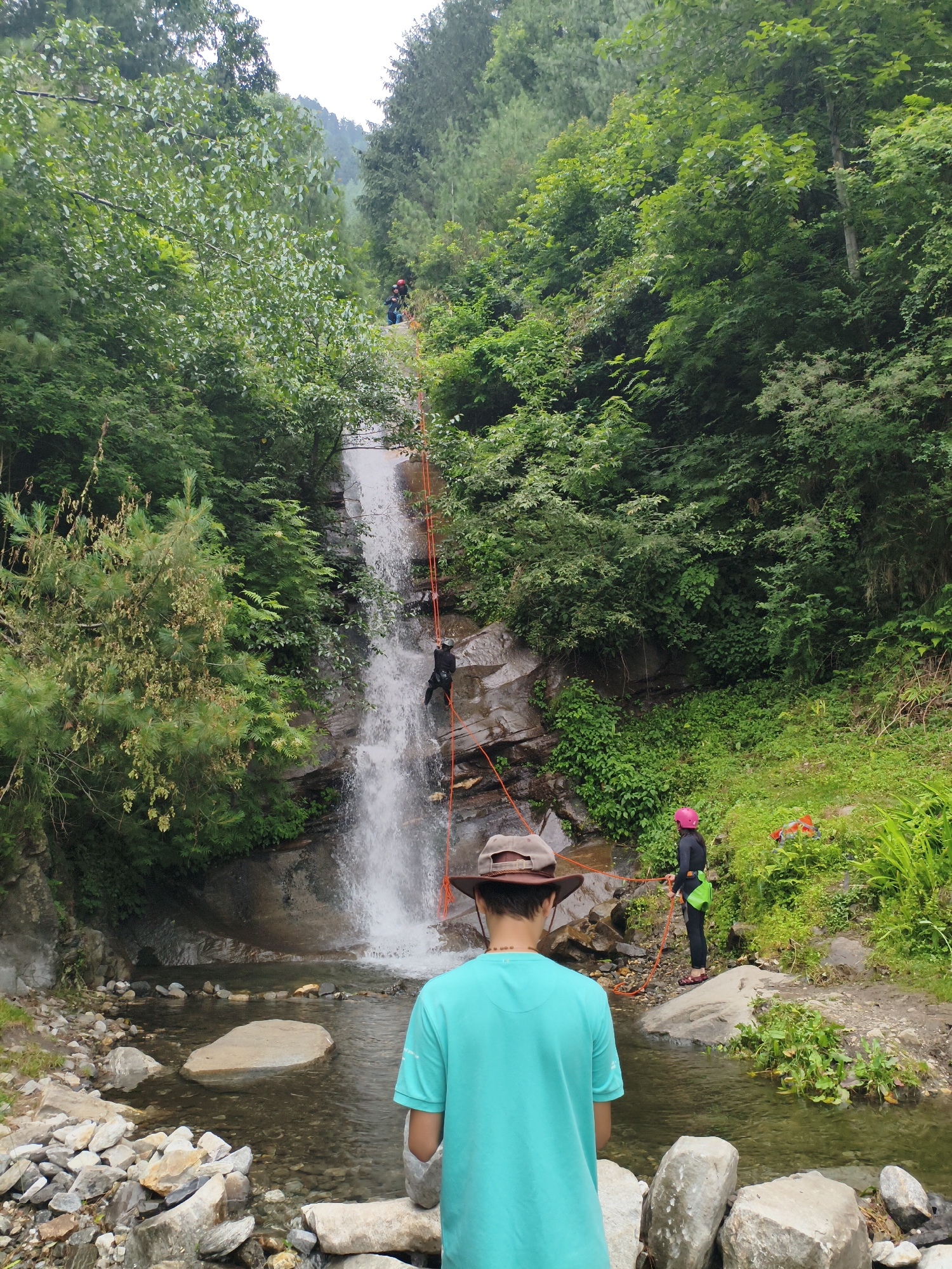 In Vashisht, India, a group of individuals engages in waterfall rappelling. One person is actively rappelling down the cascading water, suspended by an orange rope. Another person, wearing a pink helmet and black attire, stands at the water's edge, holding a rope. A third person, seen from behind, wears a teal shirt and a brown hat, observing the activity. The environment is lush and green, characterized by a rocky waterfall, a clear stream at its base, and dense foliage covering the surrounding hills.