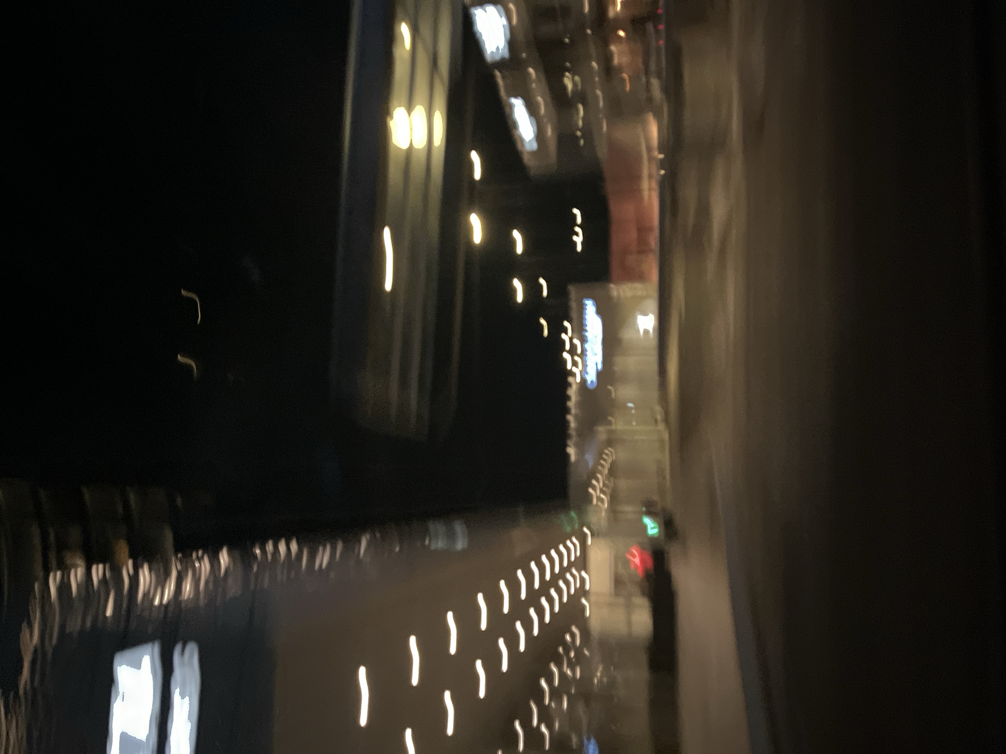 The image depicts a blurry night scene, likely taken from a moving vehicle in Puebla, Mexico. The illumination primarily consists of diffuse, elongated streaks of light, likely from streetlights and vehicle headlights. A building façade is reflected on the left side of the image, suggesting a wet surface. A series of consistent, shorter light streaks line the side of the road, indicating streetlights or reflectors. A green and red light, resembling a traffic signal, can be seen in the distance. The overall blur suggests motion and low light conditions.
