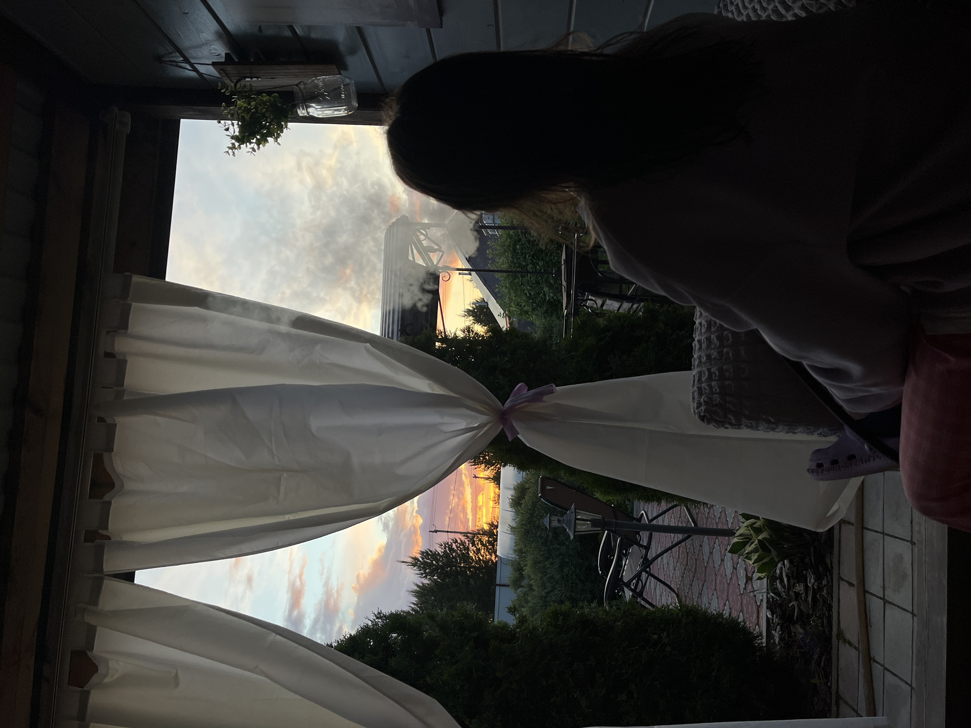 A person sits within a structure resembling a gazebo, viewing an outdoor scene. The scene is framed by white curtains tied back with a lilac ribbon. Beyond the curtains, there are evergreens. Visible behind them, the sky contains clouds and orange light, likely due to sunrise or sunset. A table and chairs are partially visible among the vegetation.
The location of Chaikovsky, Russia is not verifiable.