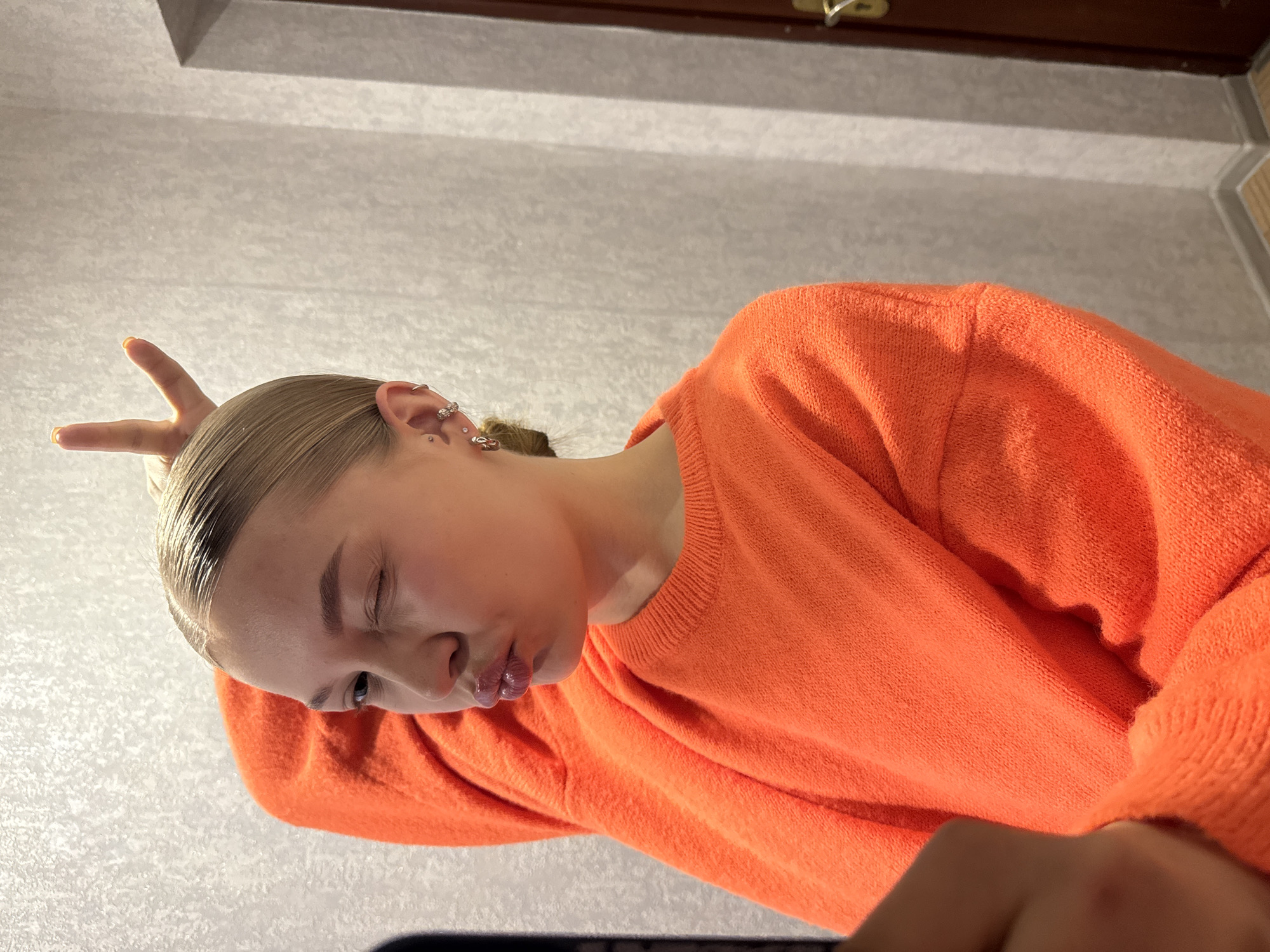 A person with light skin and slicked-back blonde hair, wearing an orange sweater, is making a bunny ear gesture with their fingers above their head. The person's face is tilted downward, with one eye partially closed and pursed lips. The background appears to be a neutral-colored wall and a dark wooden structure, possibly a headboard or furniture detail. There is no contextual information to indicate the location is in Chaikovsky, Russia.