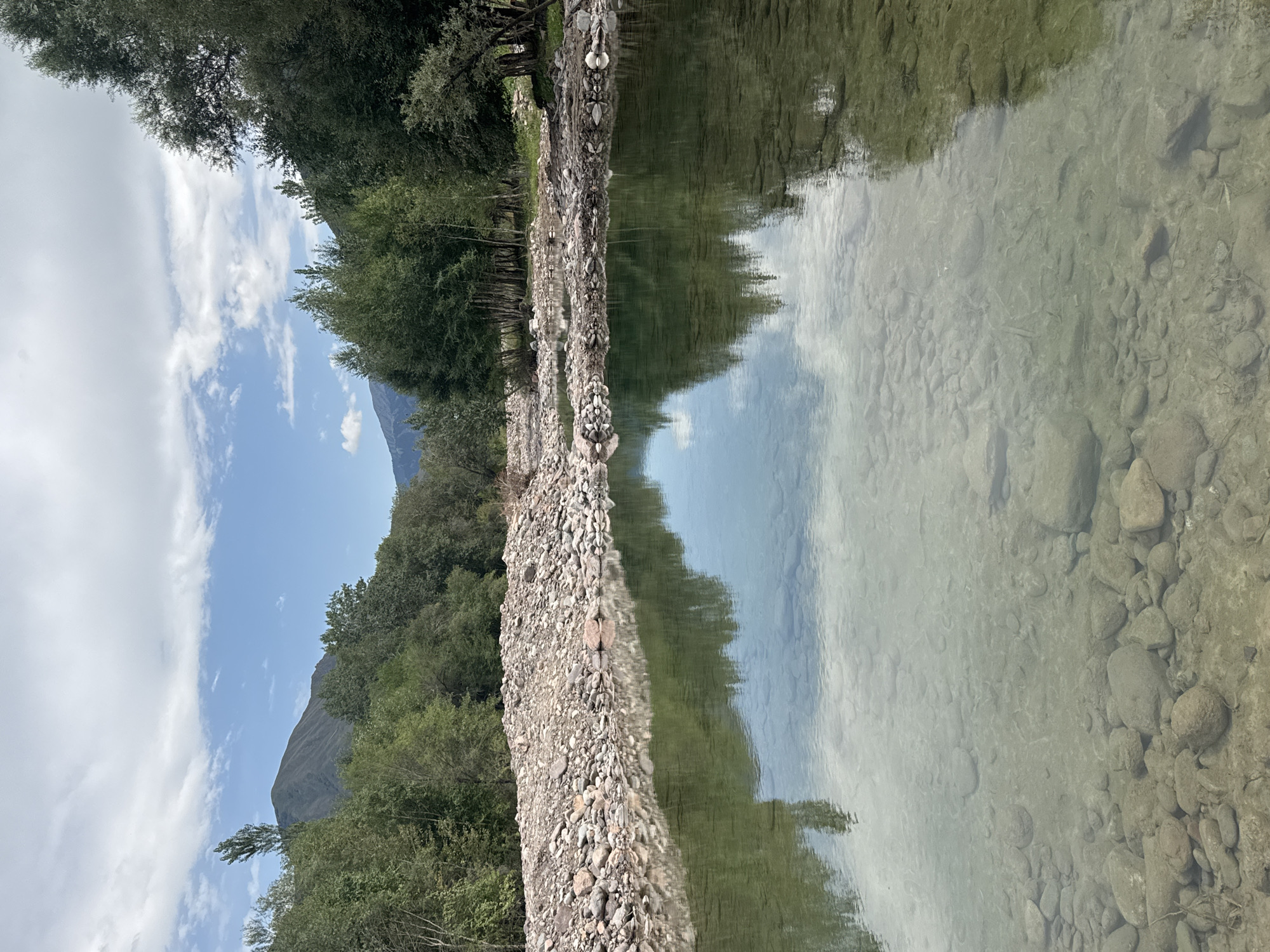 The image shows a wide, shallow river with a rocky bottom and clear water. The riverbanks are covered with stones and pebbles, and dense green trees line the opposite bank. In the background, mountains rise, partially obscured by trees and a cloudy sky. The water reflects the trees and the sky. The scene appears to be a natural landscape. The location is the river in Leskhoz, Kazakhstan.