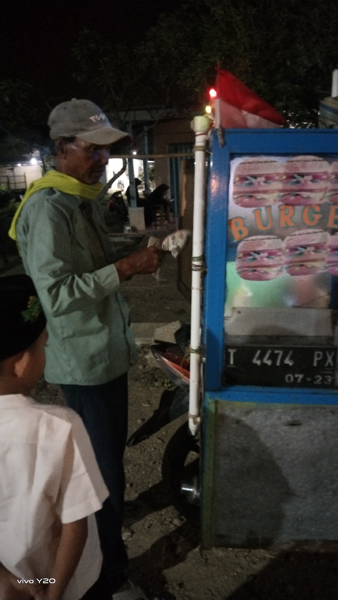 An outdoor night scene captures a man in a light blue button-down shirt, dark pants, and a grey baseball cap, standing next to a mobile food cart. He is wearing a yellow scarf around his neck and holds a white paper or cloth in his right hand. The food cart, painted blue and with a silver-colored front panel, displays images of burgers on its front. A license plate on the cart reads "T 4474 PX 07-23". A red and white flag is visible on top of the cart, along with two small red lights and a yellow light. A child wearing a white shirt and a black cap is partially visible on the left side of the image. The ground is covered in gravel and dirt. In the background, trees and structures suggest a semi-urban environment, possibly in the city of Karawang, Indonesia.