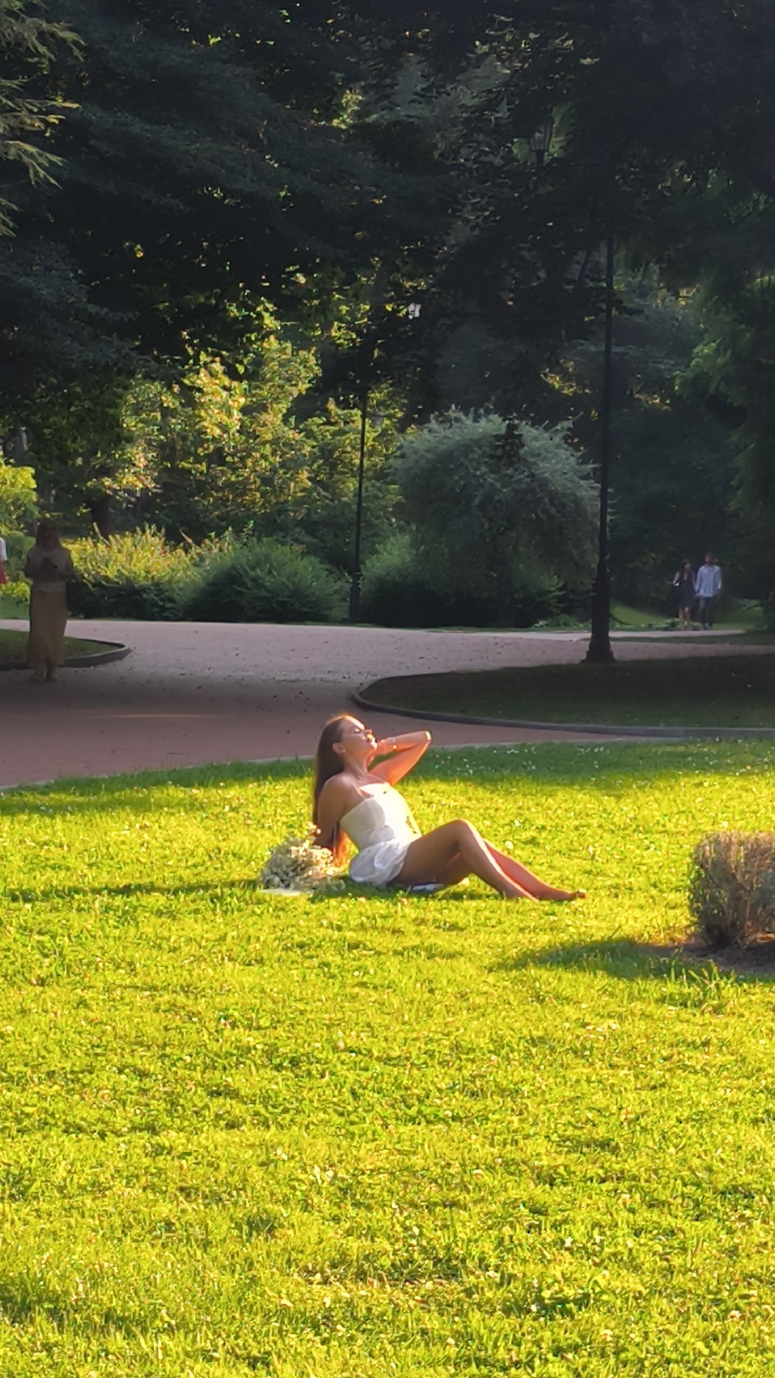 A young woman in a white dress is sitting on a grassy lawn in a park. She is reclining with her legs extended and her head tilted back, her hand resting on the back of her head. A bouquet of white flowers is beside her. The park appears to be well-maintained with mature trees in the background and a paved pathway. The sunlight casts a warm glow on the scene. In the background, two people are walking on the pathway, and another person is standing near a bush. The setting is consistent with a park in Lviv, Ukraine, possibly Stryiskyi Park, known for its scenic beauty and recreational spaces.