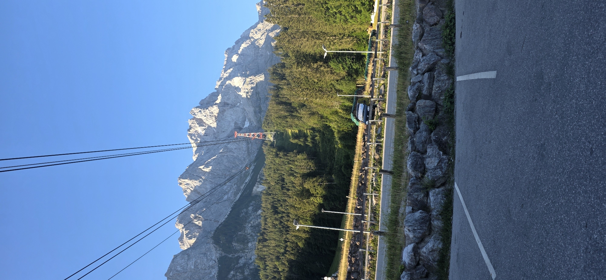 The image shows a view looking up towards a mountain range in Grainau, Germany, with a cable car system visible. In the foreground, there is a paved parking lot with white lines marking parking spaces. A low wall made of large grey rocks borders the parking lot. Behind the wall, there is a road with a wooden fence running alongside it. Beyond the road, there is a grassy area with two tall streetlights. A dark-colored truck with a green tarp covering its load is parked on the side of the road. Further up the slope, a dense forest of pine trees covers the mountainside. A red and white pylon for the cable car system stands prominently on the slope, with cables extending upwards towards the mountain peaks. The mountains themselves are rugged and rocky, with some patches of snow or lighter-colored rock visible. The sky is a clear, bright blue.