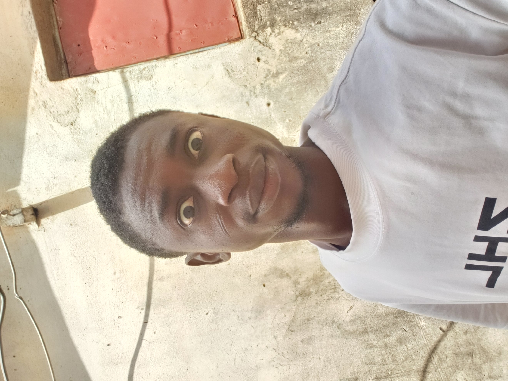 A young Black man is depicted in a close-up portrait, looking directly at the camera. He has short, dark, textured hair and a light beard or stubble. His eyes are wide, and he has a subtle, closed-mouth smile, conveying a playful or slightly surprised expression. He is wearing a white crew-neck t-shirt. On the right side of his chest, partially visible black block letters can be seen, which appear to read "HT" with an inverted character (possibly an "S" or "V") preceding them.
The background consists of a textured, light beige or off-white wall, likely stucco or concrete, showing some signs of wear and minor discoloration. In the upper left portion of the background, a reddish-brown surface or structure is visible, possibly part of a window frame or building facade. The scene is brightly lit by natural sunlight, casting soft shadows and highlighting the textures of the wall and the man's features. The setting is outdoors, in a casual, likely urban or semi-urban environment in Adankolo, Nigeria, with the bright lighting suggesting it is daytime. The man is posing for the photograph.
