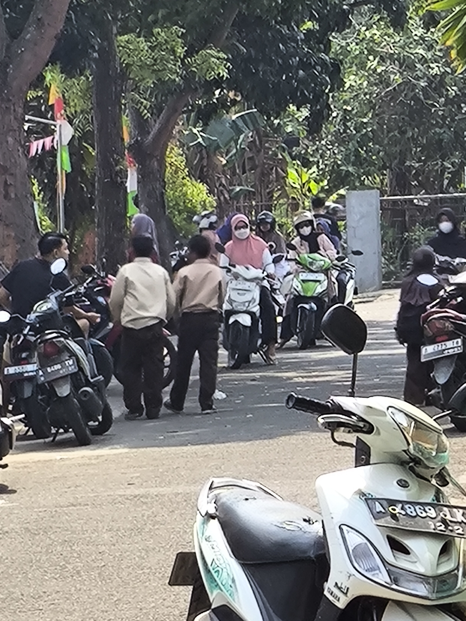 In Cilegon, Indonesia, on a sunny day, a group of individuals are gathered around several parked motorcycles. Many are wearing masks and some are wearing hijabs, suggesting the ongoing presence of public health measures. Two men, one in a light-colored shirt and dark pants, and another in a dark shirt, stand facing away from the camera near parked motorcycles. In the background, more people are seen on or around motorcycles, some wearing helmets and masks. Flags, possibly for a celebration or event, are visible in the trees. The scene appears to be on a paved road or street, with trees lining the background.