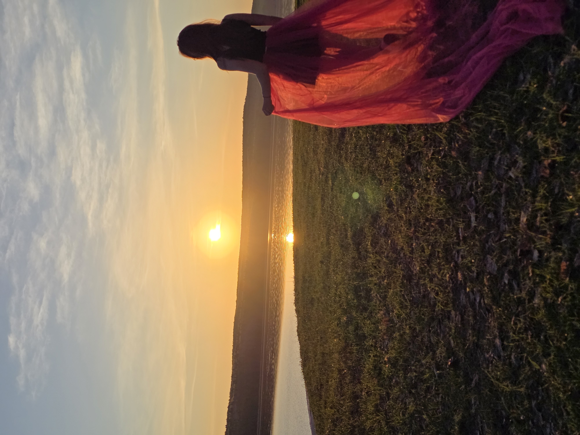 A person stands on a grassy field next to a body of water at sunset. The person is partially silhouetted and is wearing a red, flowing garment. The sun is setting over the water in the distance. The location is Chaikovsky, Russia.