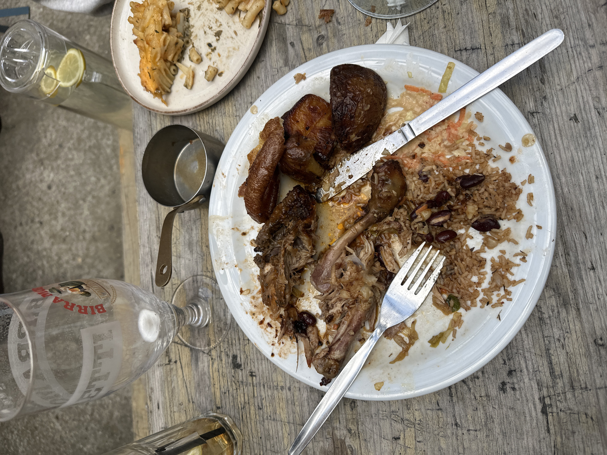 The image shows a partially eaten meal on a wooden surface. The meal includes rice with kidney beans, browned meat (possibly chicken and potatoes), and a small amount of coleslaw. There is a fork and knife on the plate. Nearby are a beer glass (branded "Birra"), a water pitcher with lemon slices, a small metal cup, and another plate with leftover pasta. The scene appears to be outdoors. There is no indication of the location being in London, UK.