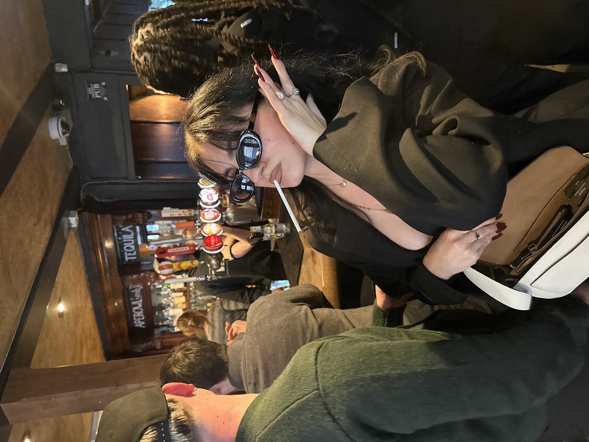 An indoor scene depicts a woman prominently in the foreground, seated at a bar counter. She has long dark hair, wears large oval sunglasses, and a black blazer. Her hands, adorned with multiple rings, are raised near her face, showcasing very long, dark red nails. A white cylindrical object, resembling a cigarette or vape pen, is held in her mouth.
The setting is a dimly lit pub or bar, characterized by wooden architectural elements and a well-stocked bar area. Visible behind the woman are several beer taps and bottles. Text on the taps includes "TEQUILA" and "APEROL SPRITZ." A sign on a black panel in the background partially reads "FRIENDLY PUB." A white security camera is mounted on a wall to the left.
Other individuals are visible in the midground and background, mostly men seated at the bar with their backs to the camera, suggesting a social gathering. One man's head, wearing a dark baseball cap, is in the immediate foreground. The woman's open handbag, cream and white in color, rests beside her, with discernible text on labels inside, including "Vogue" and "Magnum." The atmosphere appears relaxed, consistent with an evening out in a pub, potentially in London, United Kingdom given the context.