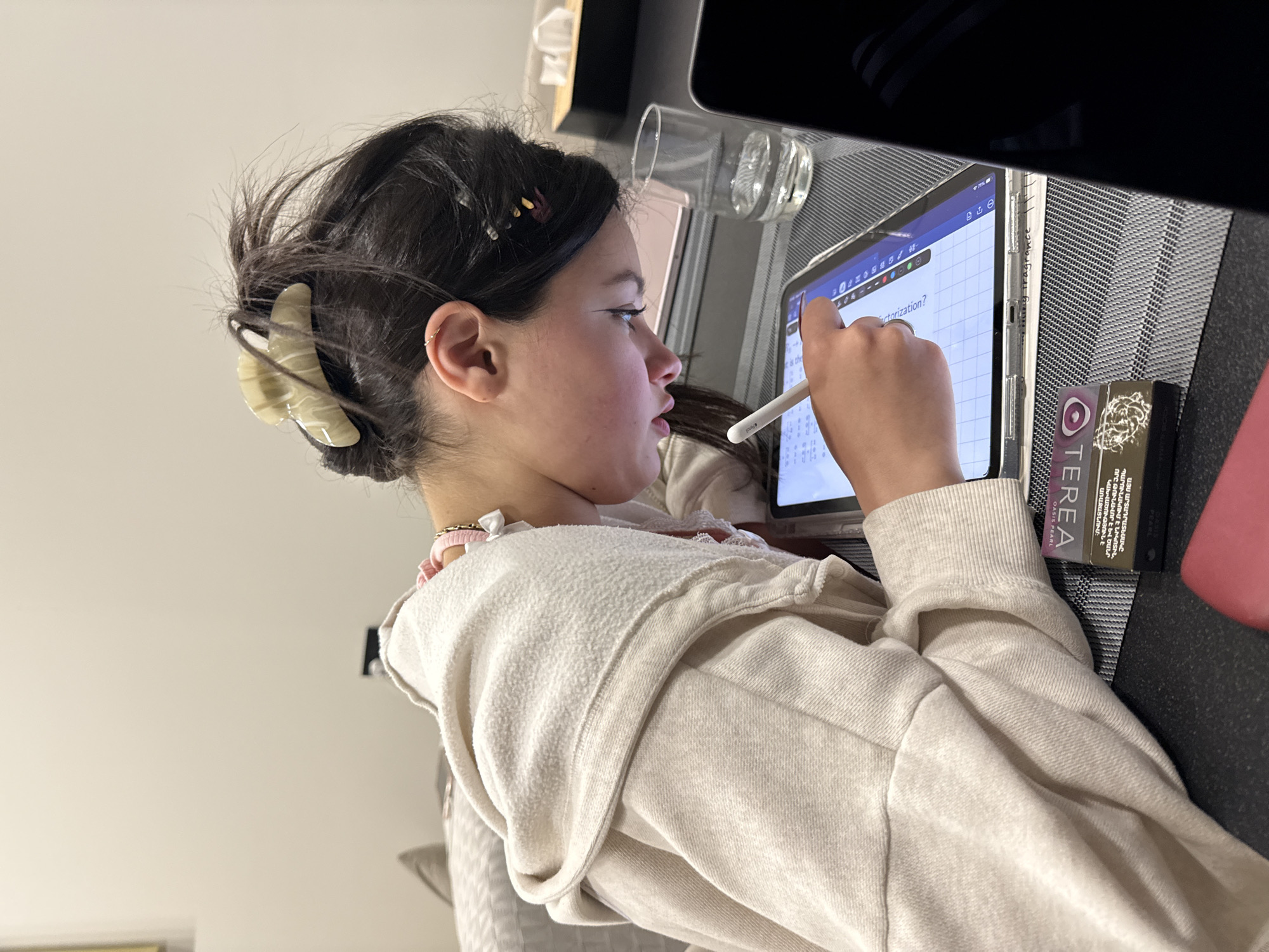 A young girl is sitting at a table working on a tablet with a stylus. She is wearing a light-colored hooded sweatshirt and has her hair pulled back with a clip. The tablet screen displays a document with text, and a box of "TEREA Oasis Pearl" smoking product is on the table next to it. The OCR indicates Armenian text. The location cannot be determined to be Yerevan, Armenia without further evidence.