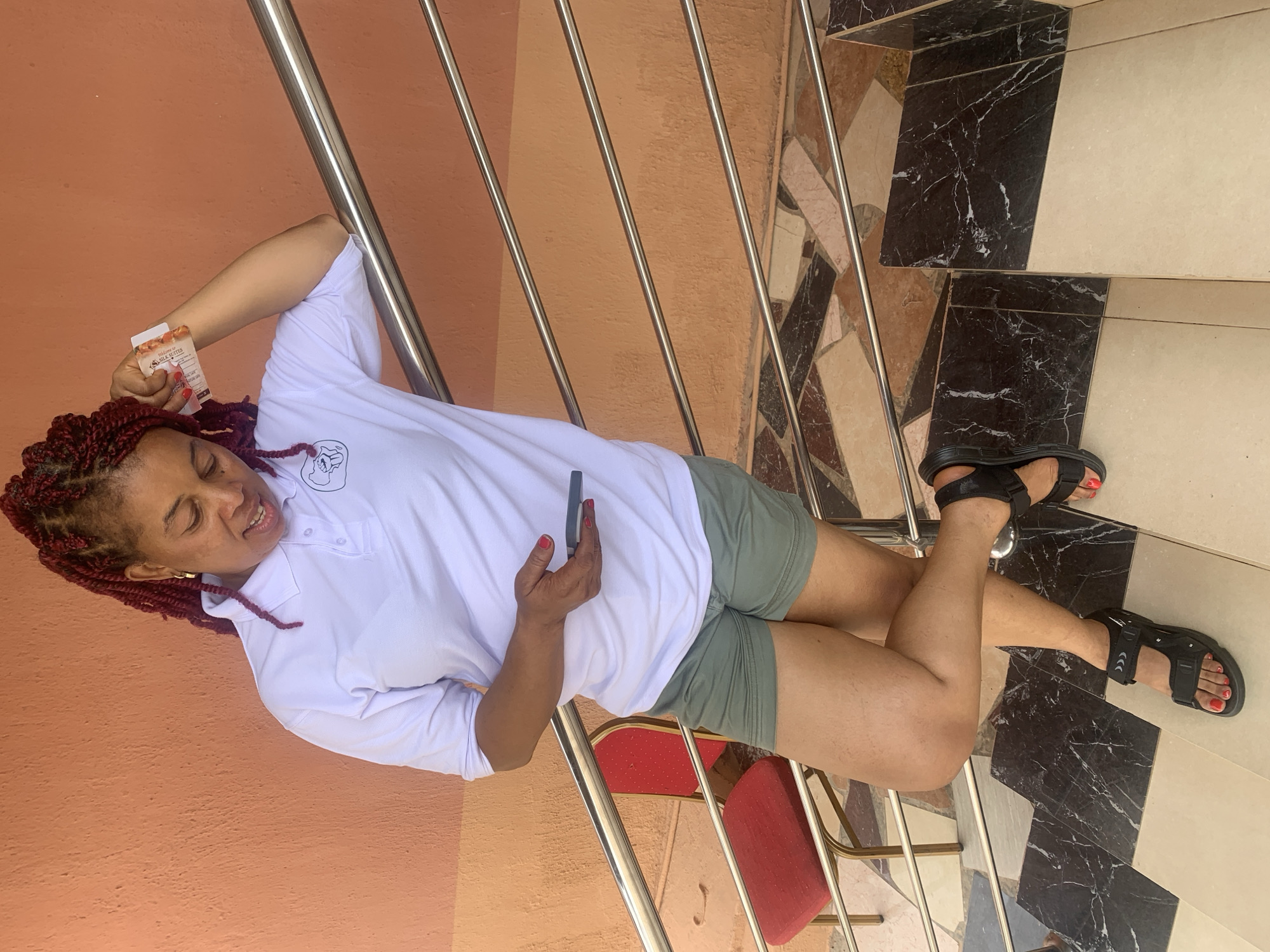 A woman with dark red braided hair, wearing a white polo shirt and green shorts, is lying on a staircase in Gura Top, Nigeria. She holds a black cell phone in her right hand and a small rectangular object in her left. Her feet are clad in black sandals. The staircase has a silver railing and the steps have a black and white marble pattern. Behind her, there is an orange wall and a red chair.