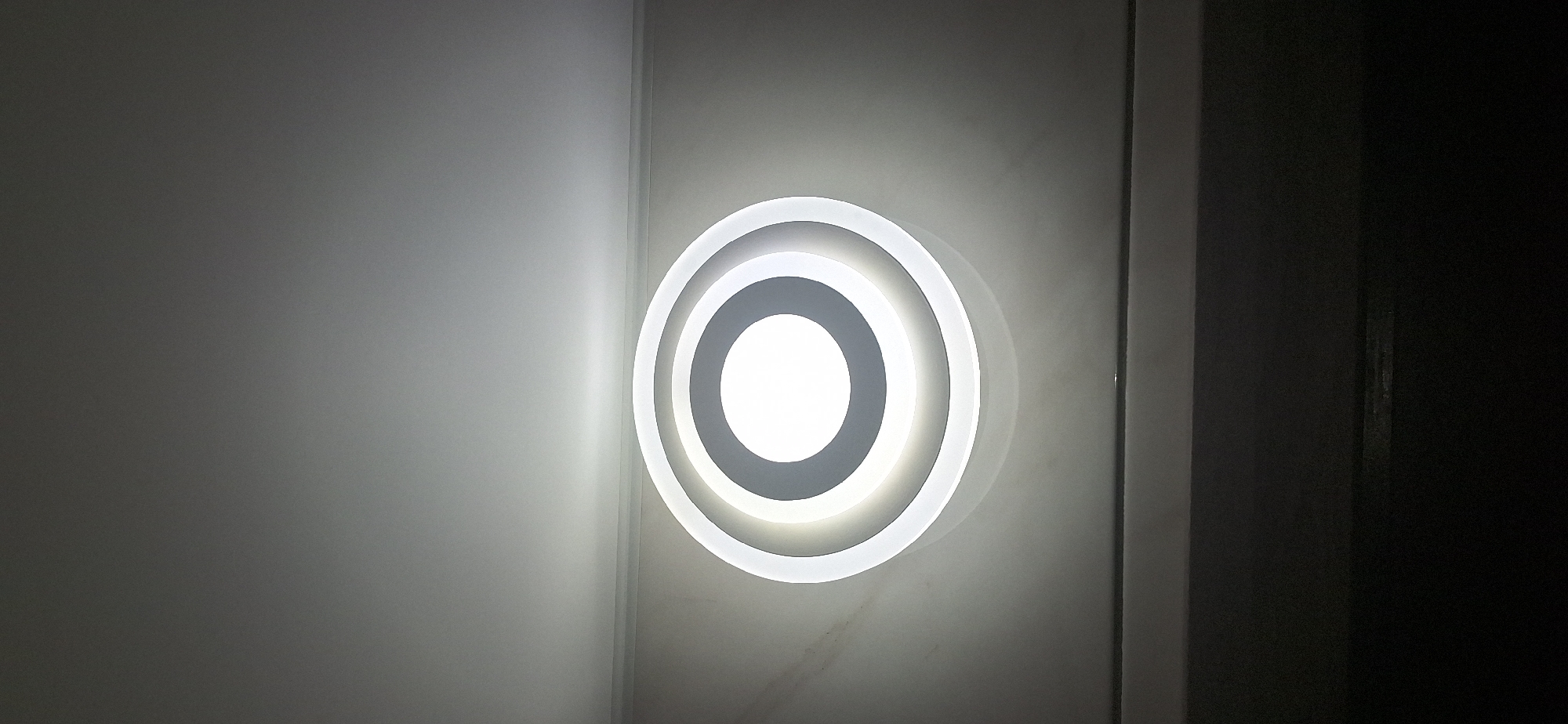 The image shows a modern, circular ceiling light fixture with concentric rings. The innermost ring is illuminated with a bright white light, creating a halo effect that extends to the outer rings. The fixture is mounted on a tiled ceiling, which appears to be a neutral color with some subtle marbling. The lower portion of the image is dark, suggesting it is a room interior with dim lighting. The image does not provide any specific details or context that would allow for identification of its location within the city of Bila Tserkva, Ukraine.