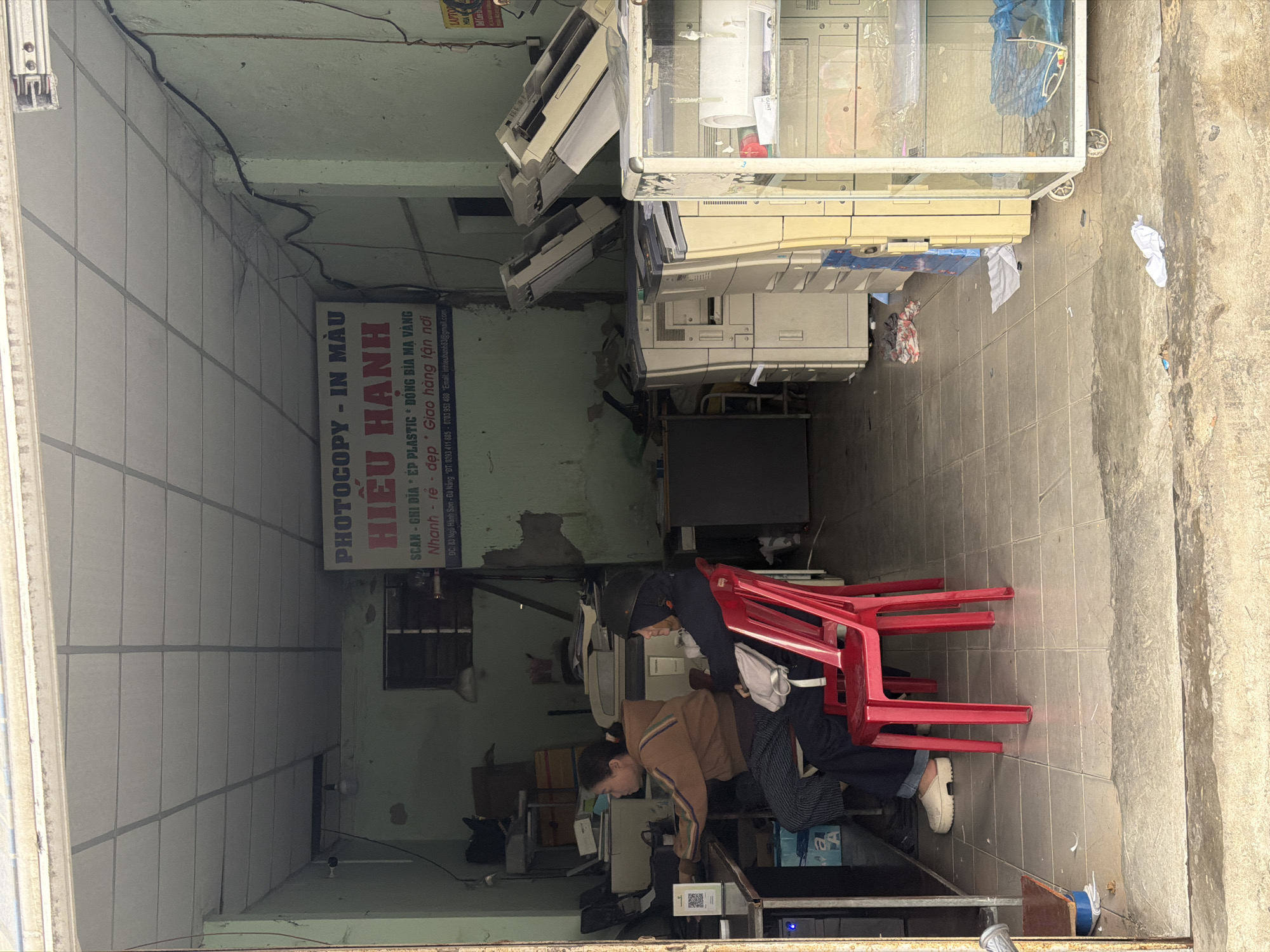 The image captures the interior of a photocopy and printing shop named "HIỆU HẠNH" in Đà Nẵng, Vietnam, viewed from just outside the entrance. The shop appears functional and somewhat cluttered, with light green walls showing some signs of wear and a tiled floor. The ceiling is made of light-colored panels with exposed wiring.
Two individuals are present in the shop. One person, wearing a brown striped top, is leaning over a counter in the foreground, seemingly engaged with equipment or tasks. A QR code sticker is visible on the counter near them. Another person is seated behind them on a red plastic chair, wearing a dark jacket and a helmet, with a white bag slung over the chair. Several other red plastic chairs are stacked nearby.
The shop is filled with various office machinery, including multiple large, beige-colored industrial photocopiers and printers, some stacked. A clear display case on wheels is positioned on the right side of the shop, containing papers and other items. Miscellaneous boxes, papers, and supplies are visible throughout the space. The ambient lighting suggests it is daytime.
A prominent sign on the back wall advertises the shop's services in Vietnamese, including:
- "PHOTOCOPY - IN MÀU" (Photocopy - Color Printing)
- "HIỆU HẠNH" (Hieu Hanh - shop name)
- "SCAN - GHI DÍA * ÉP PLASTIC * ĐÓNG BÌA MẠ VÀNG" (Scan - Write CD/DVD * Plastic Laminating * Gold-plated Binding)
- "Nhanh - rẻ - đẹp * Giao hàng tận nơi" (Fast - Cheap - Beautiful * Delivery to location)
- Contact information: "ĐC: 83 Ngũ Hành Sơn - Đà Năng *ĐT: 0393 411 885 - 0703 953 480 *Email: inhieuhanh83@gmail.com"