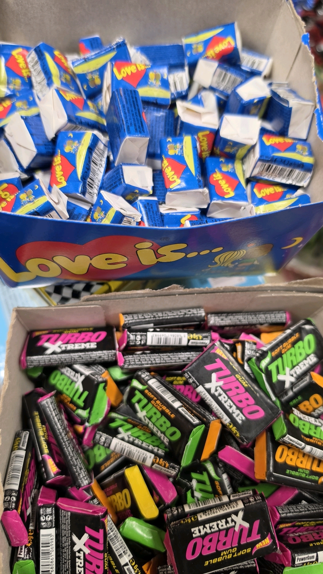The image shows two open cardboard boxes filled with individually wrapped chewing gum. The top box contains "Love is..." brand chewing gum, with blue and yellow wrappers featuring a red heart and cartoon illustrations. The bottom box contains "Turbo Xtreme" brand chewing gum, with black wrappers that have pink and green lettering. Some of the "Turbo Xtreme" wrappers also have checkered patterns. The lighting suggests an indoor setting, possibly a shop or storage area. There are no people visible in the image. The overall scene depicts a collection of confectionery products. No information is available regarding its location in Jūrmala, Latvia.