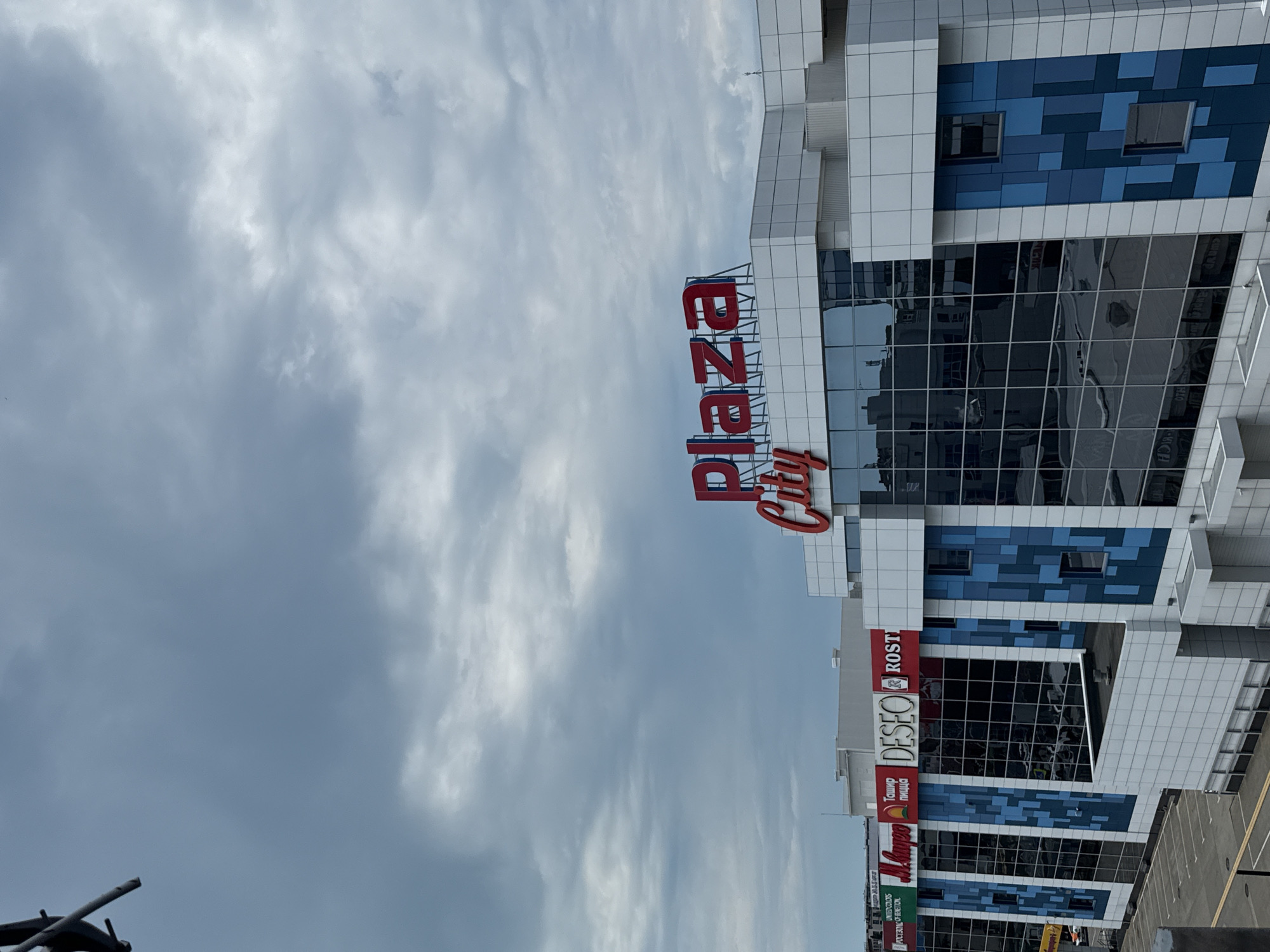 The image shows a contemporary shopping center in Sochi, Russia, on a cloudy day. The building features a modern design with a white exterior and large glass windows. Several signs are visible on the facade, including "Plaza City" in large red lettering at the top. Below this, various store names are displayed, such as "M.Video", "Тащир пицца", "DESEO", and "ROST". The sky is overcast with heavy, gray clouds. The foreground shows a parking lot with parking spaces marked. The overall impression is that of a functional, urban commercial space.