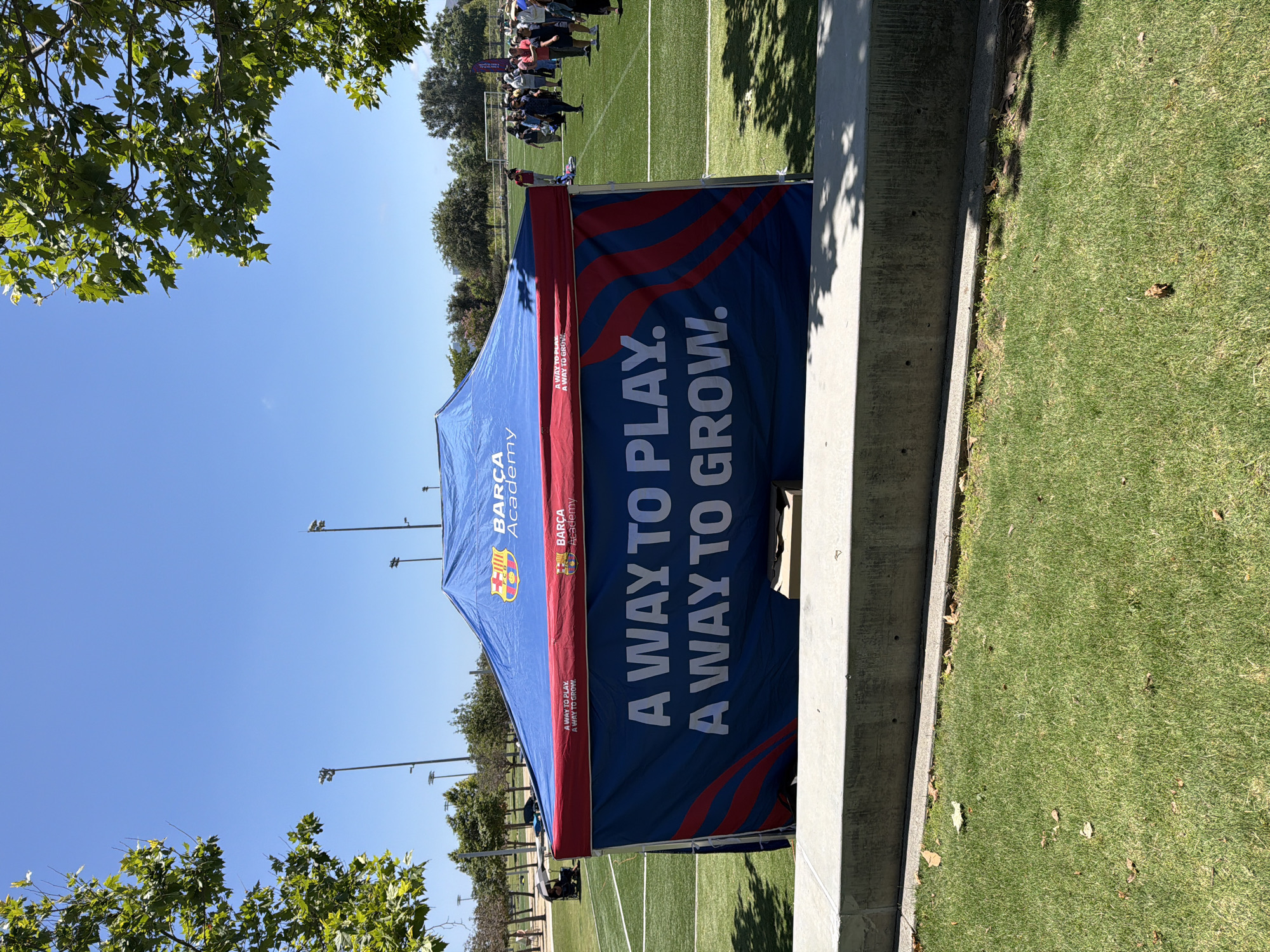 A Barcelona Academy tent is set up on a sunny day on a green turf field in Irvine, United States of America. The tent is blue with red trim and features the Barcelona Academy logo and the text "A WAY TO PLAY. A WAY TO GROW." in large white letters. Several people are gathered in the background, some standing and some sitting, suggesting an event or activity is taking place. The field is marked with white lines, typical of a sports field, and stadium lights are visible in the distance. Trees with green foliage frame the top of the image, casting shadows on the tent. The foreground shows a concrete barrier and a grassy area.