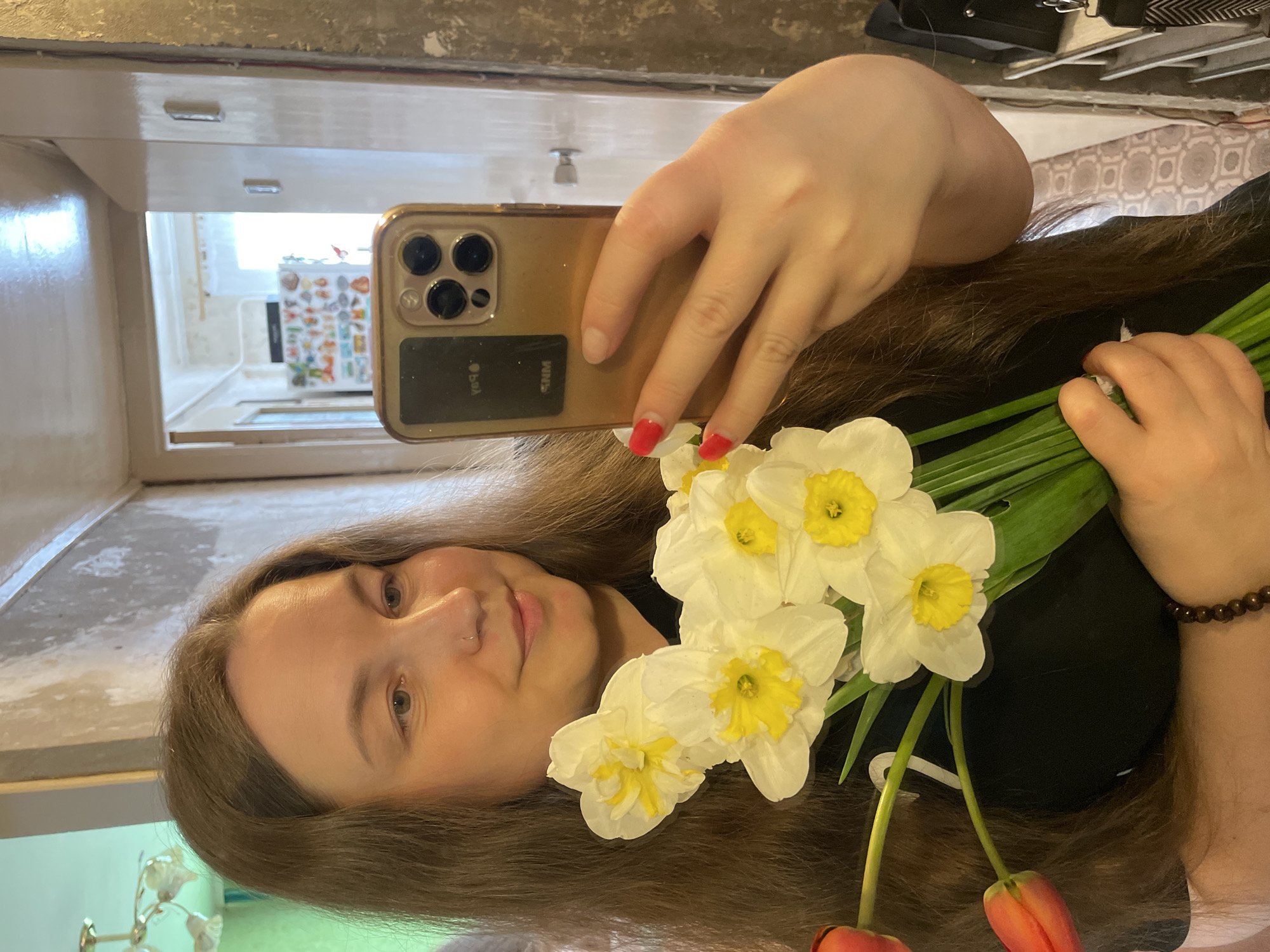A woman is holding a bouquet of white and yellow daffodils and red tulips in front of herself, taking a selfie with a gold-colored iPhone. She has long brown hair and is wearing a black shirt. Her nails are painted red. The background appears to be a hallway, visible in a mirror behind her. The reflection in the mirror reveals a white door frame and a glimpse of a room with a refrigerator covered in magnets or stickers. The precise location within Izhevsk, Russia, cannot be determined from the provided image.