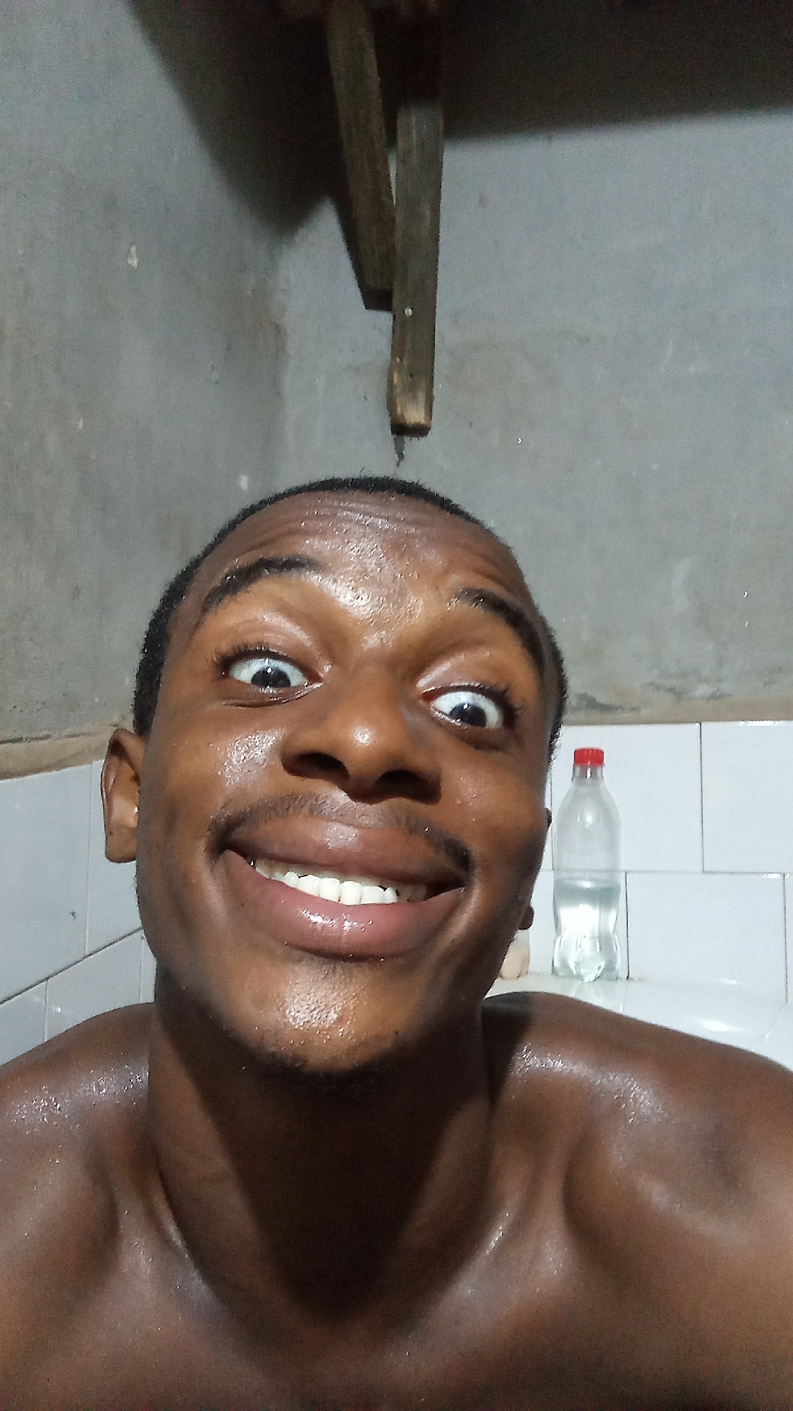 A close-up selfie captures a young Black man from the shoulders up, featuring a highly expressive and playful facial gesture. He gazes directly at the camera with wide, bulging eyes and a broad, open-mouthed smile, revealing a full set of white teeth. His skin, including his face, shoulders, and chest, appears wet or sweaty and glistening, suggesting he may have just showered, exercised, or is in a humid environment. He has short, dark hair and a faint mustache.
The setting appears to be an indoor space, likely a bathroom or a utilitarian room. The upper part of the background consists of a rough, light gray wall, possibly concrete or plaster. Below this, behind his right shoulder and extending to the right of his head, there is a section of white tiled wall. A clear plastic water bottle with a red cap, partially filled with water, sits on what appears to be the edge of a sink or bathtub on the tiled surface. Above the man's head, an aged, rustic wooden structure, possibly consisting of planks or a small shelf, is visible against the gray wall. The lighting seems artificial and somewhat dim. There is no visible text in the image. The man's exaggerated expression conveys a sense of humor and energy.