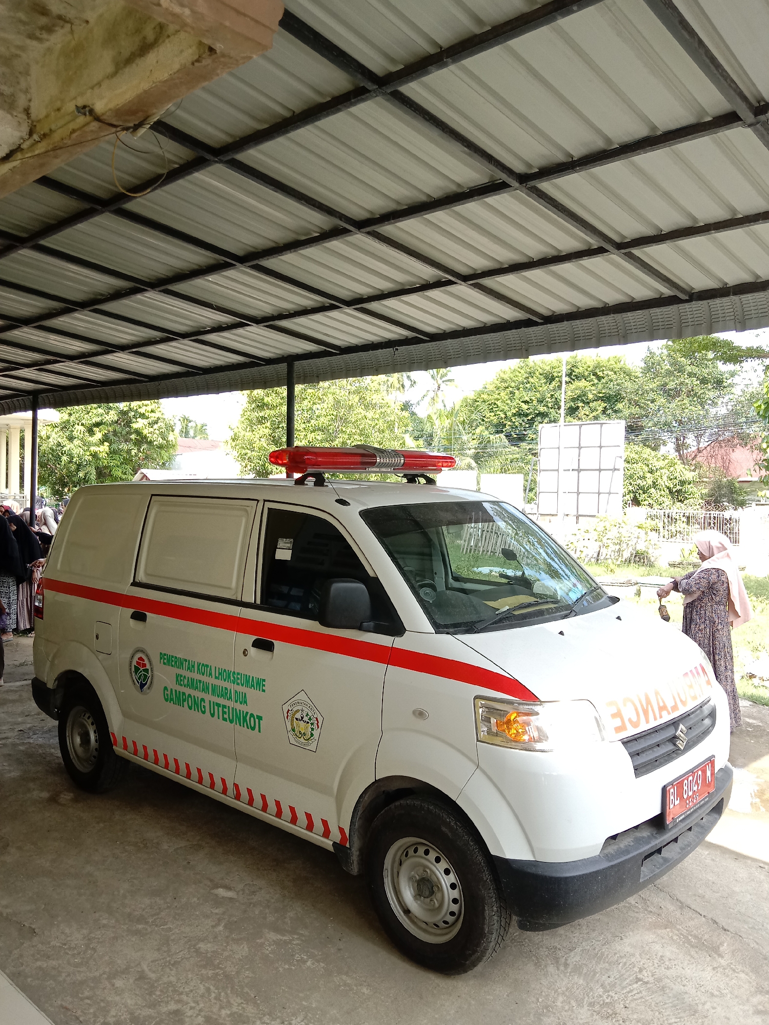 The image shows a white ambulance parked under a metal awning. The ambulance has red stripes and emergency lights on top. The words "PEMERINTAH KOTA LHOKSEUMAWE KECAMATAN MUARA DUA GAMPONG UTEUNKOT" are printed on the side of the vehicle, identifying it as belonging to the local government of Lhokseumawe, specifically the Muara Dua subdistrict, Uteunkot village, Indonesia. A woman wearing a headscarf is standing near the front of the ambulance, seemingly interacting with it. Other people are visible in the background to the left. The environment consists of a partially covered area, likely a carport or similar structure, and greenery in the background. The license plate of the ambulance is visible, reading "BL 8049 N".