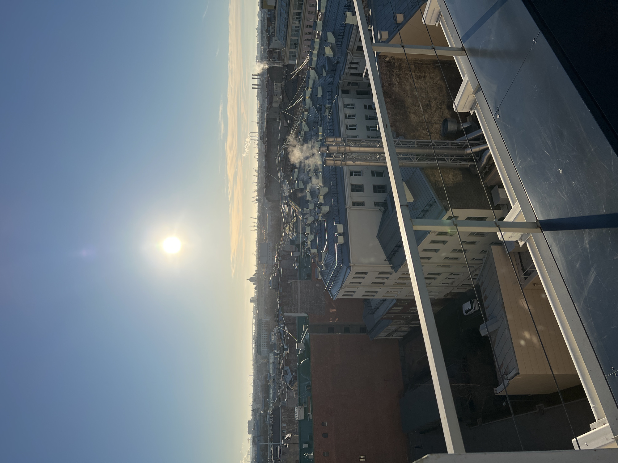 The image shows an elevated view of Saint Petersburg, Russia, likely taken from a tall building. The sky is blue with the sun visible. Buildings with various roof types and colors are visible in the middle ground. Some structures appear to have industrial components like smokestacks with smoke plumes. The foreground contains elements of the structure the image was taken from, including reflective glass panels and supporting metal framework.