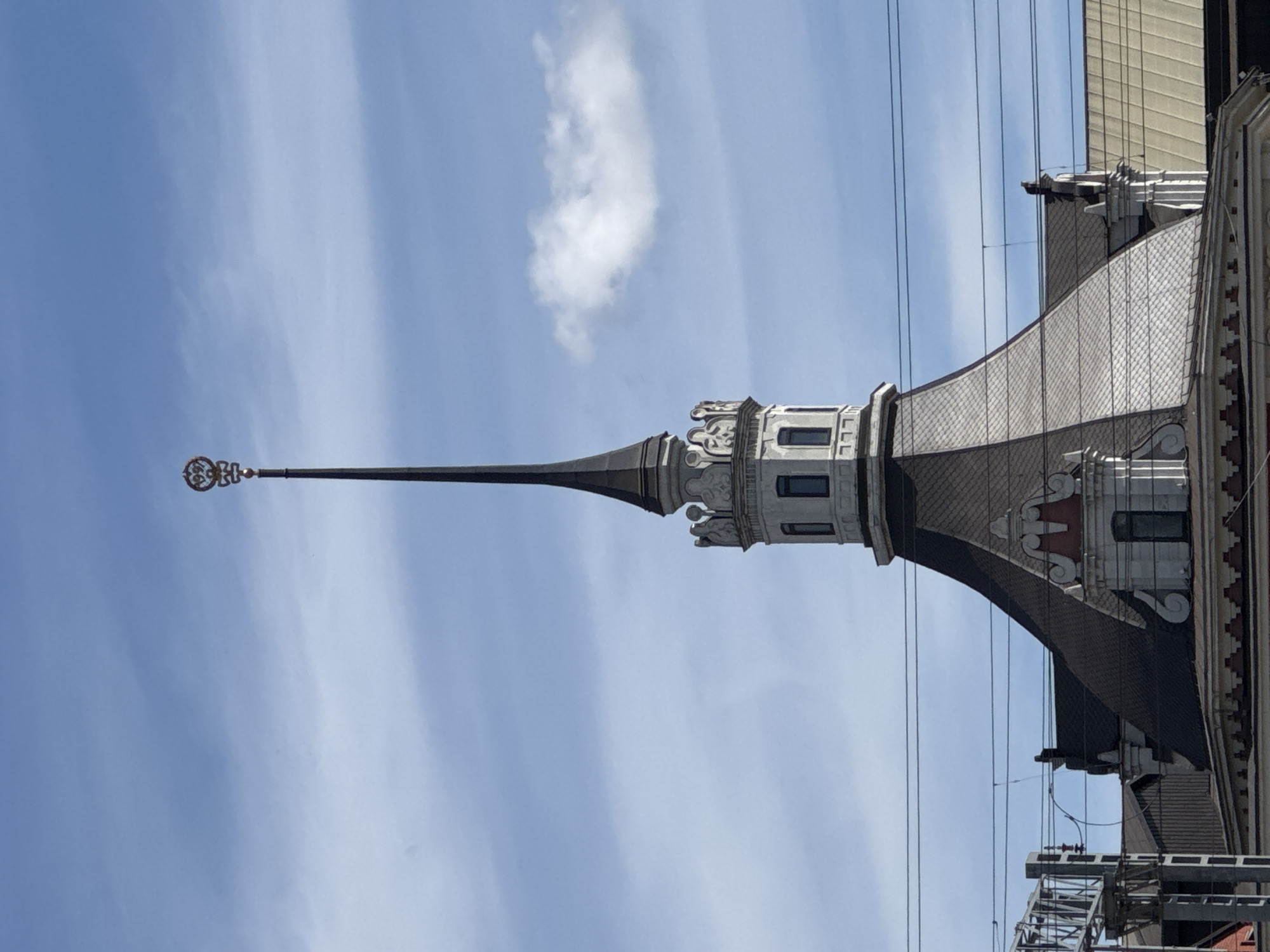 This media file, oriented vertically, captures a detail of a building's spire against a bright, slightly cloudy sky. The spire is tall and slender, tapering to a fine point adorned with an intricate, circular, gold-colored finial. The spire's base is part of a more elaborate architectural feature with decorative stonework, including carved gargoyles or grotesques, and visible window frames. The roofline below the spire is covered in grey, textured tiles, creating a curved silhouette. In the background, thin, dark power lines crisscross the sky, suggesting an urban setting with infrastructure. The bright lighting and the presence of a single, wispy cloud indicate it is daytime, likely late morning or early afternoon, under fair weather conditions. There is no visible text in the image. The image is focused on architectural details and the sky, evoking a sense of height and historical architecture within a modern, developed environment. The vertical orientation of the image emphasizes the upward reach of the spire.