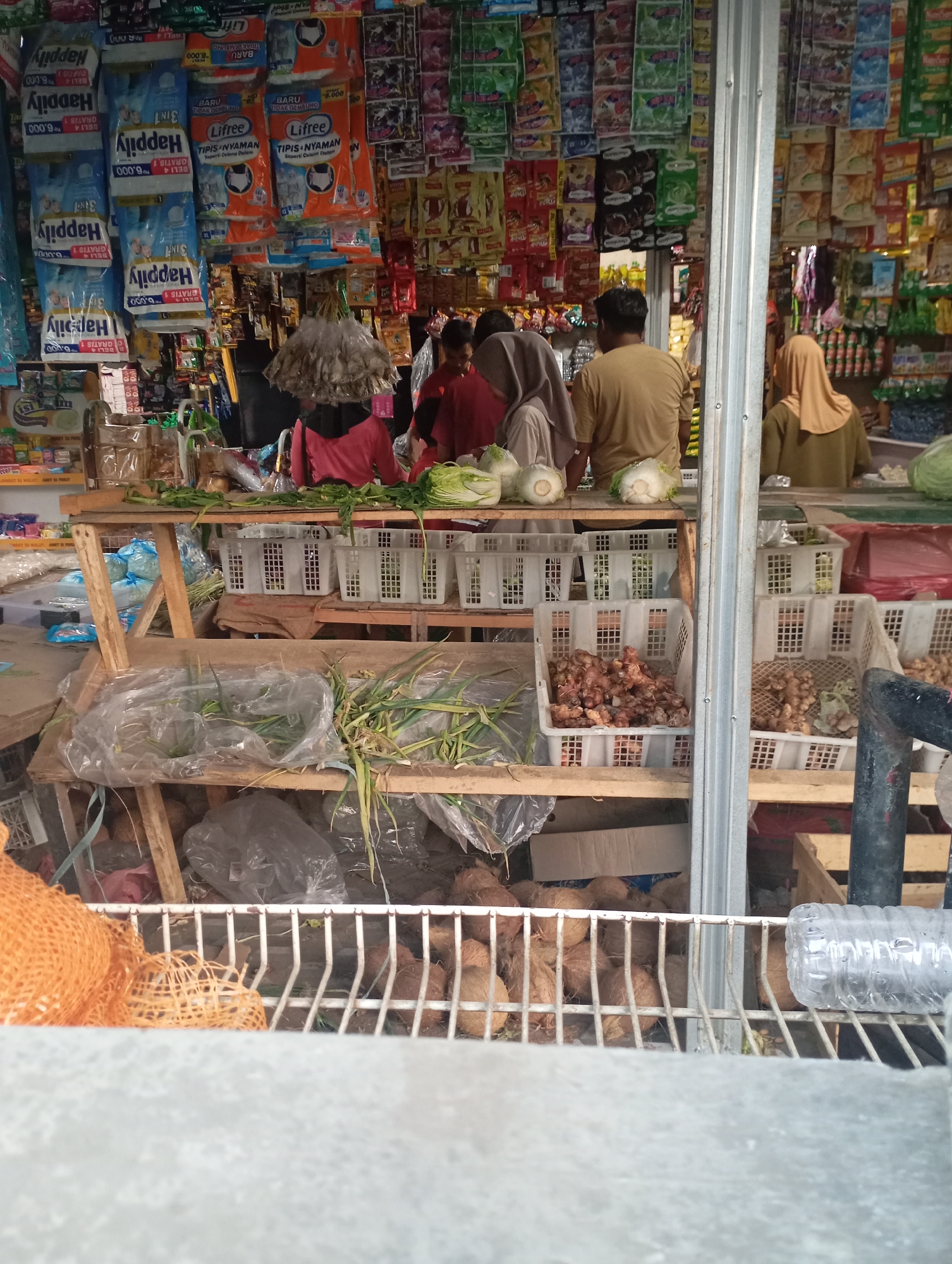 This image captures a street-side market stall in Kecamatan Haurgeulis, Indonesia. The stall is densely packed with various goods. In the foreground, a metal rack displays coconuts and an orange net bag, suggesting a variety of produce. Behind this, a wooden shelving unit holds plastic-wrapped vegetables like scallions, along with white plastic crates filled with ginger and other root vegetables. Further back, several people, including women wearing headscarves and a man in a tan shirt, are engaged in activities within the stall, possibly as vendors or customers. The background is filled with shelves and hanging displays of packaged goods, ranging from detergents to food items, typical of a small Indonesian convenience store or "warung." The overall impression is of a bustling, functional marketplace.