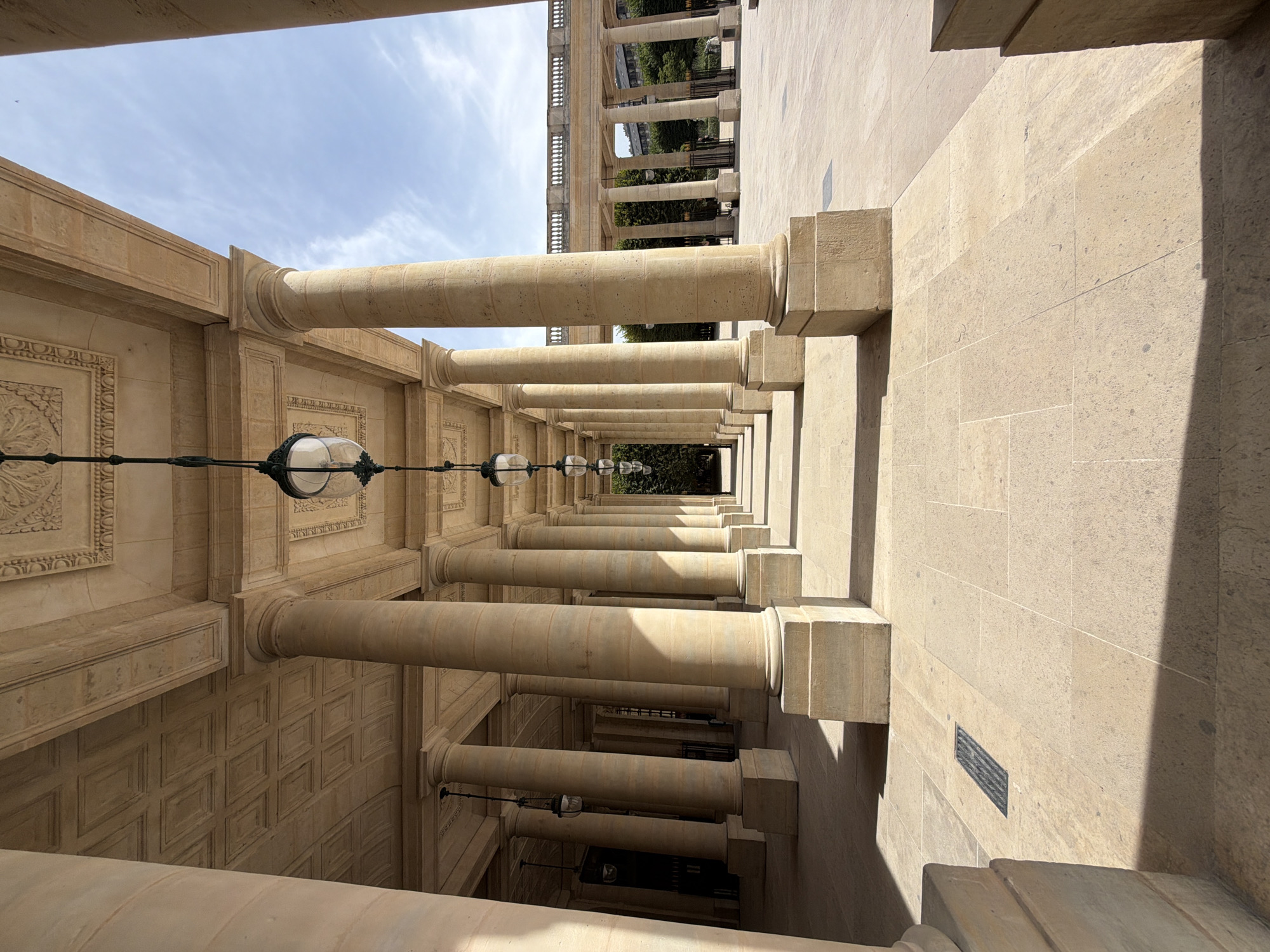 This is a low-angle, eye-level, full shot of a colonnade with several rows of tall, light-beige stone columns supporting an ornate, coffered stone ceiling. The colonnade extends into the distance, creating a strong sense of perspective. Several vintage-style hanging lanterns are suspended from the ceiling. The ground is paved with large, light-beige stone tiles, and shadows from the columns and ceiling are cast across the pavement. In the background, there is a clear blue sky with scattered white clouds. Beyond the colonnade, to the right, a balustrade and more columns of a similar architectural style are visible, with trees and distant buildings in the far background. The scene appears to be a public square or courtyard in Paris, France, likely the Palais Royal. There are no people clearly visible in the frame.