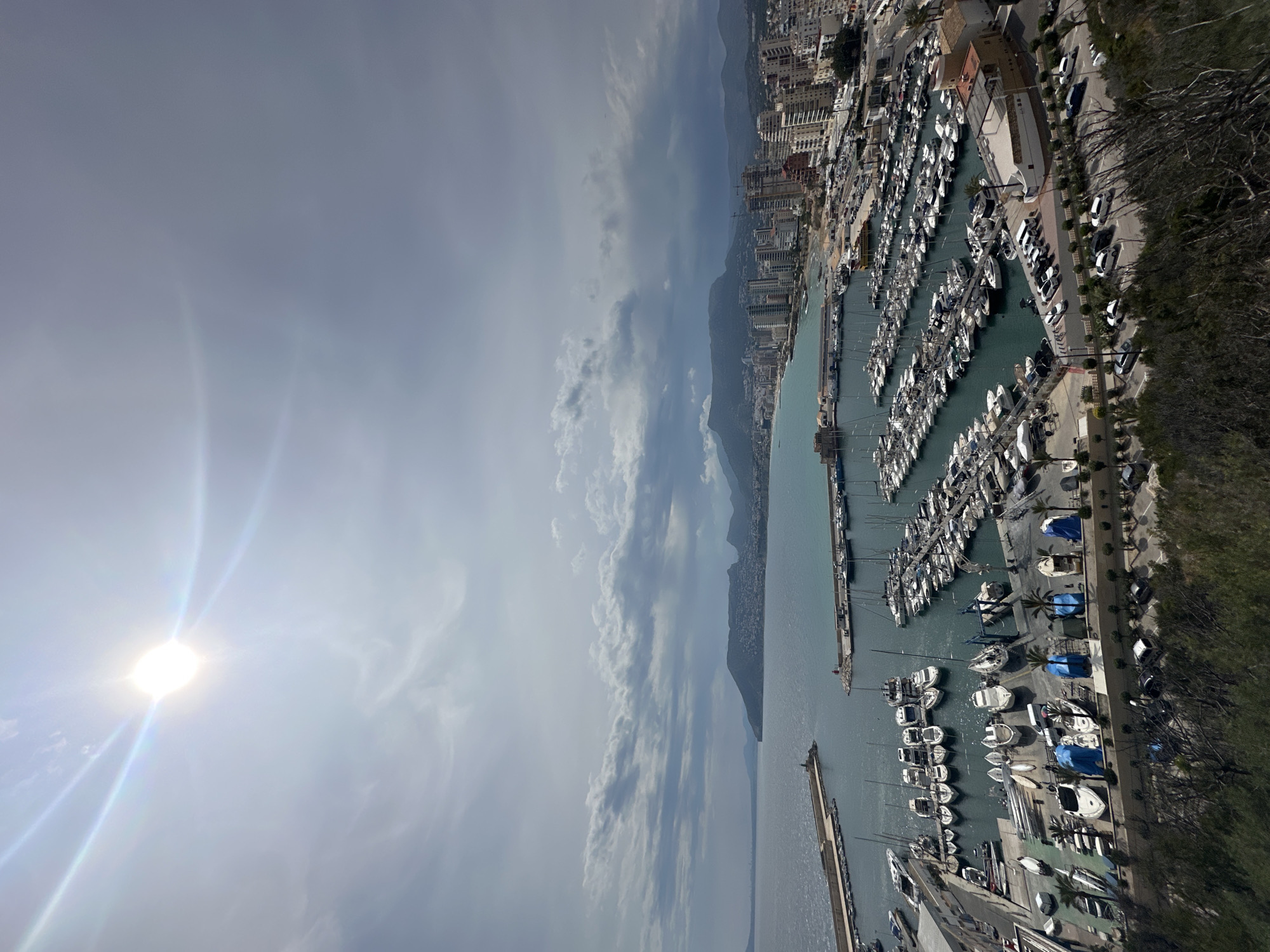 The image shows an aerial view of the marina in San Gabriel, Spain, on a sunny day with some cloud cover. Numerous boats of varying sizes are docked in organized rows within the marina's harbor. Buildings and other infrastructure line the coastline, suggesting a densely populated urban area. The water is calm, and the overall scene depicts a vibrant coastal environment.