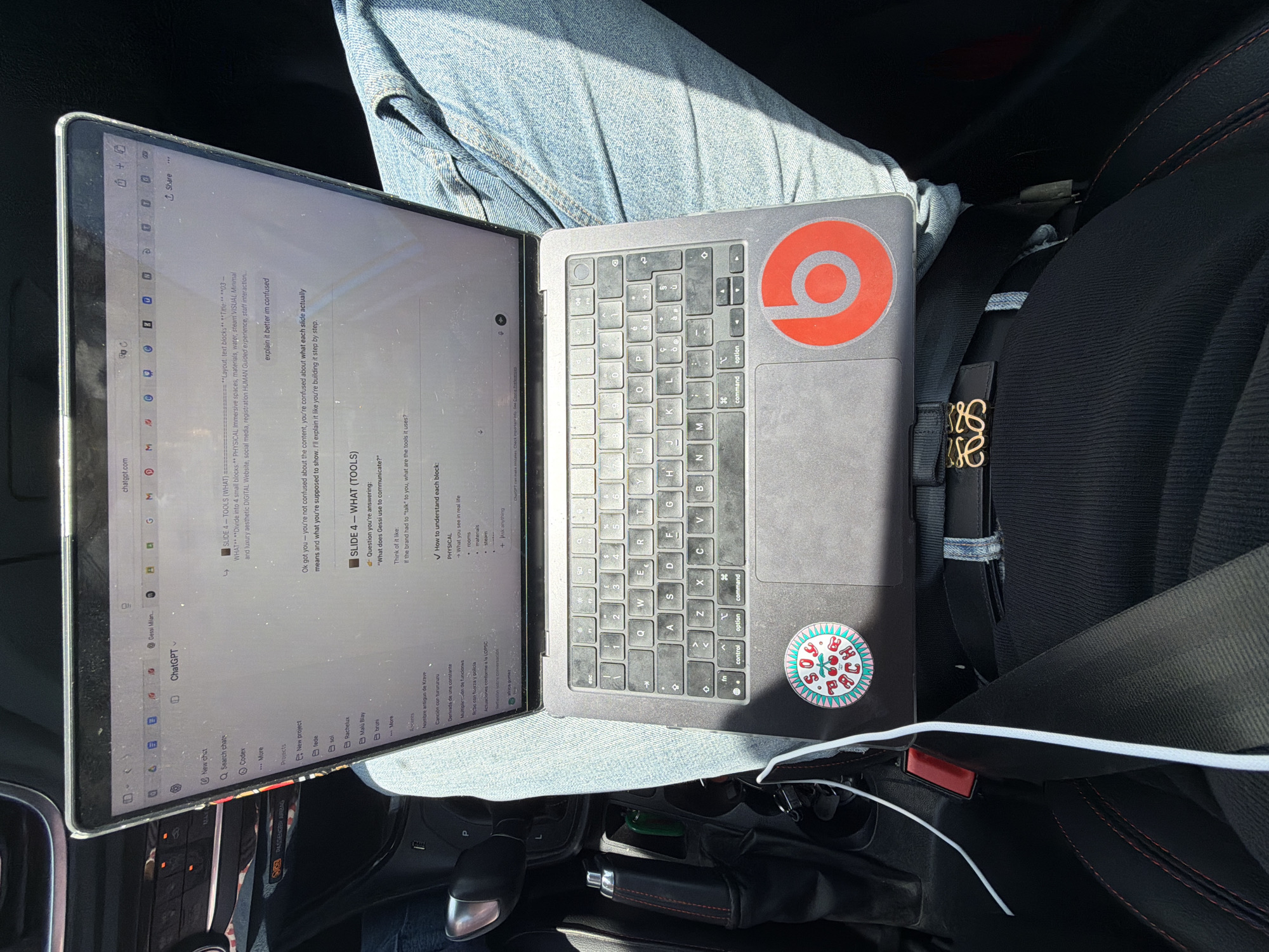 A MacBook laptop rests on a person's lap, covered by blue jeans, inside the passenger seat of a car. The bright sunlight entering from the left indicates it is daytime with sunny weather. The car's interior features black upholstery with red stitching on the seat and gear shifter boot. A black strap with a gold "LOEWE" logo, likely from a bag, is visible secured by the seatbelt. A white charging cable runs along the car's console.
The laptop screen displays a web browser with "chatgpt.com" open, showing a conversation related to a presentation or project. The text "SLIDE 4 - TOOLS (WHAT)" is visible, along with bullet points detailing "PHYSICAL Immersive spaces, materials, water, steam VISUAL Minimal and luxury aesthetic DIGITAL Website, social media, registration HUMAN Guided experience, staff interaction..." The chat interface includes prompts like "explain it better im confused" and a response beginning "Ok got you - you're not confused about the content..." Other visible text includes "New chat," "Q Search chats," "Projects," and a list of names. A warning label on the dashboard reads "SO PASSENGER AIRBAG."
The laptop's lid features a red "Beats" logo sticker, and the palm rest has a round, colorful sticker. The keyboard appears well-used. The setting suggests someone is working or browsing while in transit or parked in Sollana, Spain.