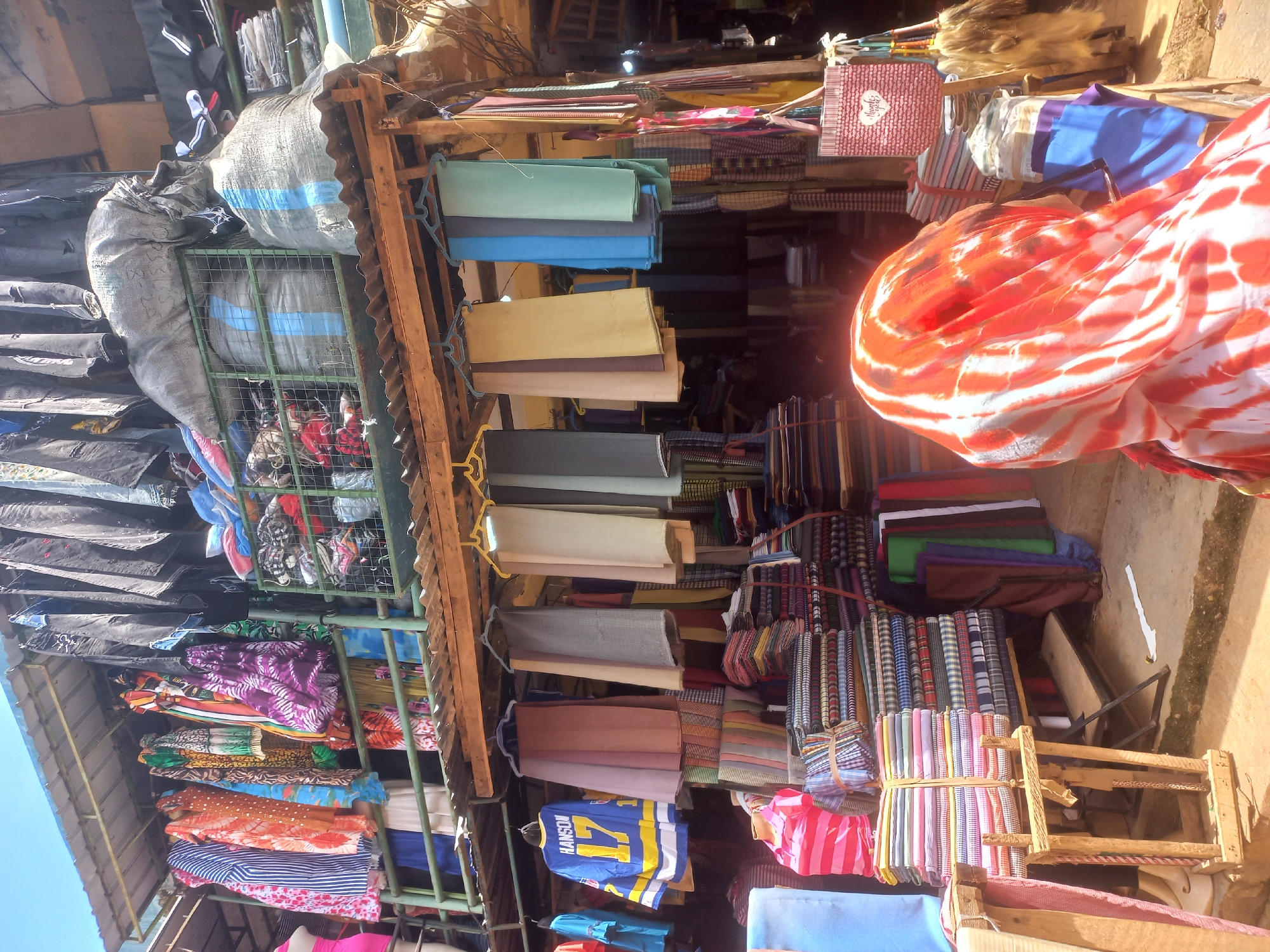 This is a photograph taken at a market in Jos, Nigeria. In the foreground, a person wearing a headscarf with an orange and white tie-dye pattern is partially visible, facing away from the camera. Behind them is a stall displaying a variety of textiles.
The stall features neatly folded stacks of patterned fabrics, with some bundles tied with string. There are also garments hanging on display, including shirts and what appear to be traditional African wear in vibrant colors and patterns. Further back, more fabrics are visible, some rolled and some folded, in various shades of blue, green, brown, and red. A mannequin in a pink and white striped shirt stands near the stacked fabrics.
Above the stall, there is a roof structure supported by wooden beams, with more clothing hanging on racks. Black and gray garments are prominently displayed overhead. Large, grey, netted bags filled with what appear to be textiles are also stored on a higher level. The overall impression is of a bustling market scene where textiles are the primary commodity. The bright daylight suggests the photo was taken during the day.