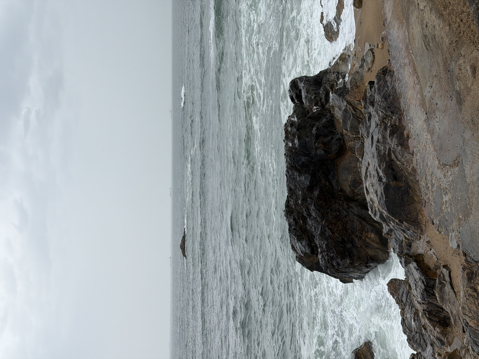 A landscape-oriented photograph displays a gray, turbulent sea meeting a rocky shoreline under an overcast sky. The photograph is oriented with the sky and sea to the left and the rock formations to the right. The sea's surface is disturbed with white-capped waves, indicating a windy or stormy condition. Several smaller rock formations protrude from the sea in the distance. A large, dark rock formation juts into the sea from the right side of the frame. It is covered with wet, dark rock, and the waves are crashing around its base. The rocky terrain extends further to the right, where it transitions into a sandy beach. The sky is uniformly gray, indicating heavy cloud cover. The horizon line is slightly tilted and blends into the sky. This image may be taken in or near Porto, Portugal, based on the appearance of the rocky coastline and the typical weather conditions of the region.
