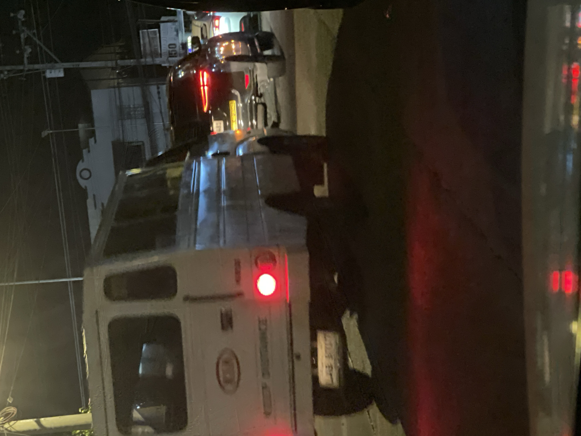 The image shows a nighttime street scene in Davao City, Philippines, likely along a main road due to the presence of multiple vehicles in traffic. The foreground is dominated by the rear of a white van, possibly a passenger van or a delivery vehicle, with its brake lights illuminated red. To the right of the van, a dark-colored SUV is visible, also with its brake lights on, indicating traffic is stopped or moving slowly. Further down the road, other vehicles and streetlights can be seen. The environment appears to be urban, with buildings and utility poles lining the street, suggesting it's a commercial or residential area. The overall impression is a typical traffic congestion scenario during nighttime hours.