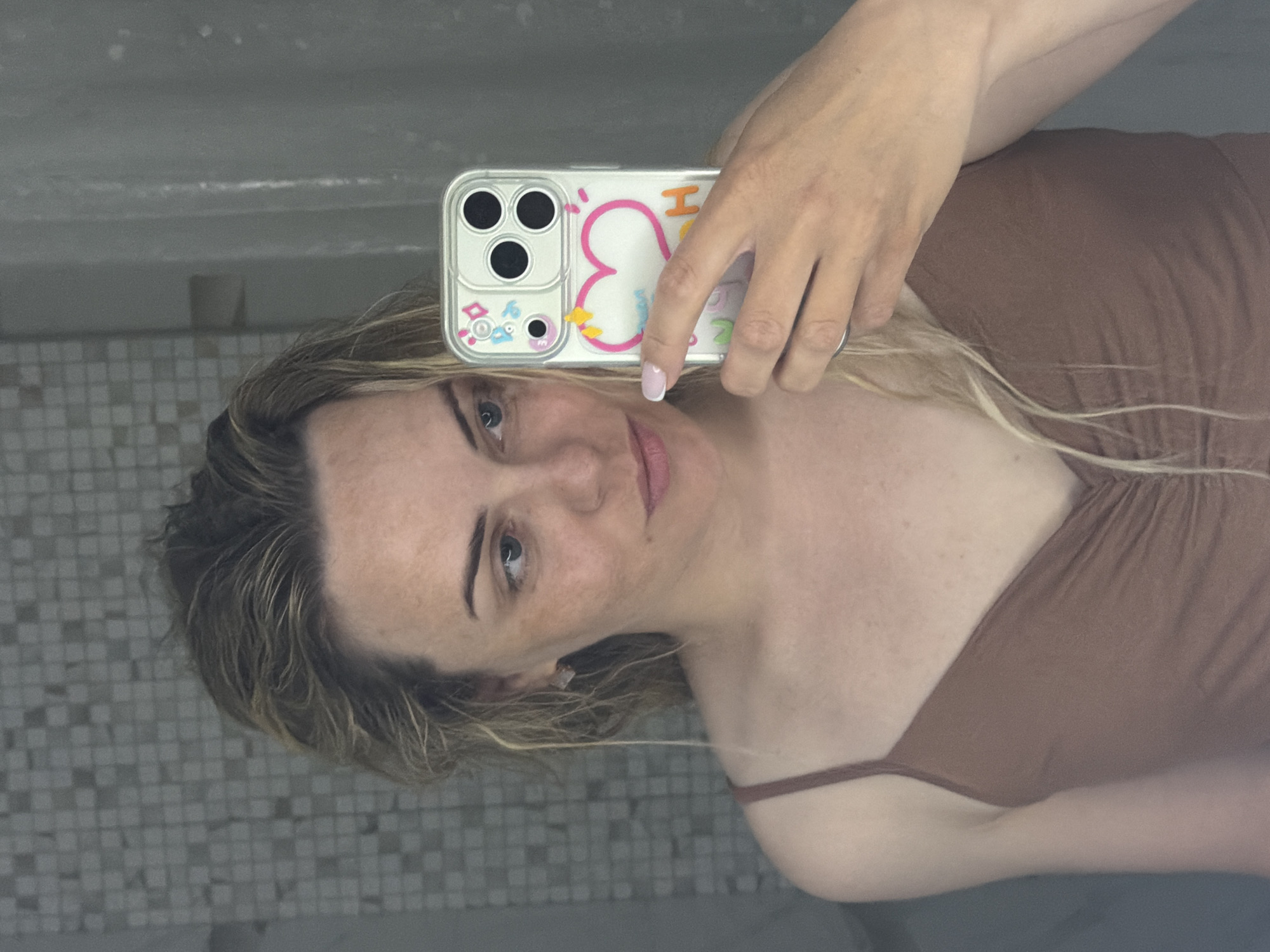 A woman is holding a cell phone, which has a decorative case, up to her face in a mirrored bathroom. The bathroom features tiled walls and a partially visible sink. The woman is wearing a brown top. The photograph's location in Votkinsk, Russia cannot be determined from the visual information provided.