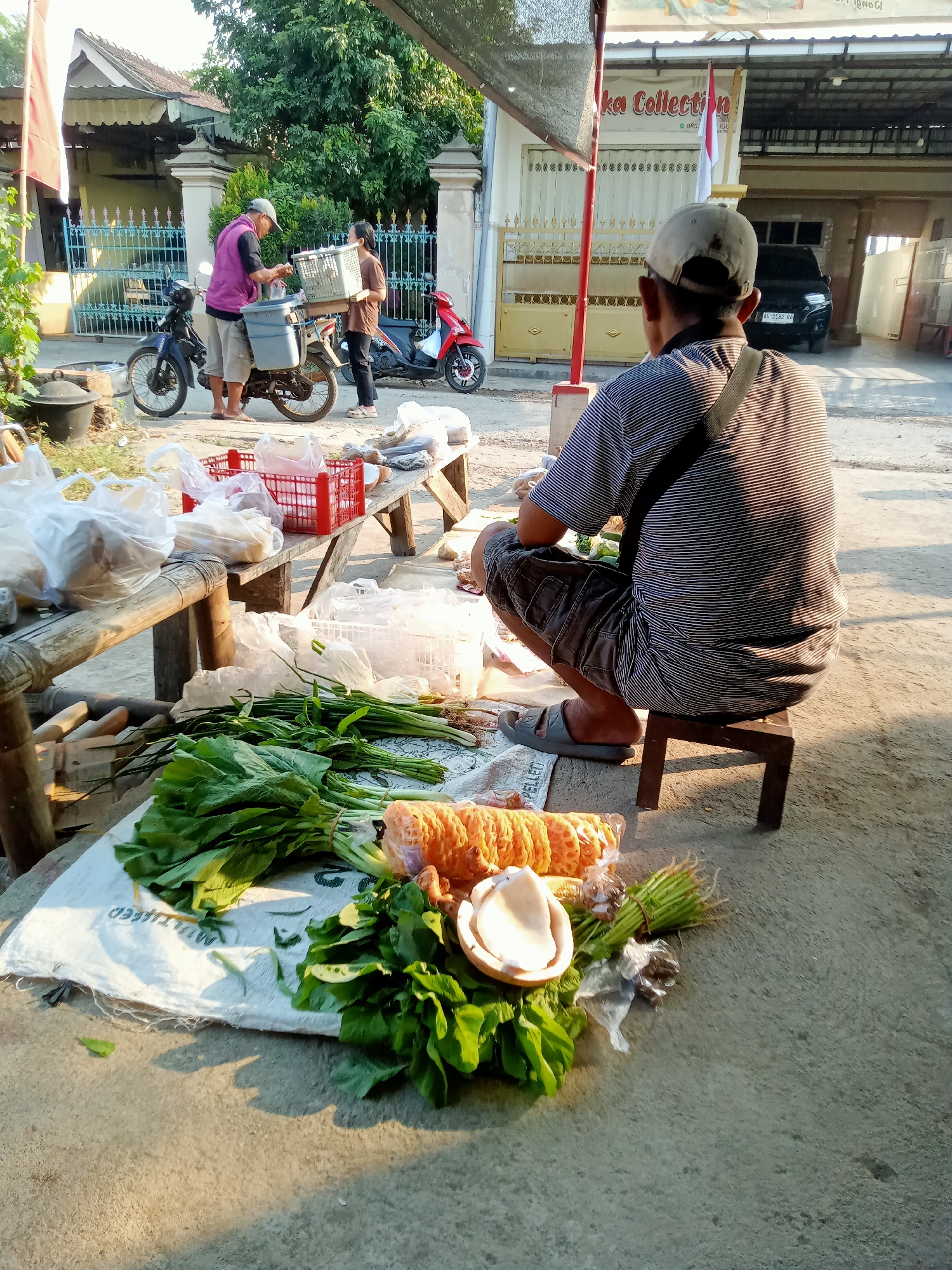 An outdoor market scene is depicted in Simo, Indonesia, under bright daylight. In the foreground, a man wearing a striped t-shirt and shorts sits on a small wooden stool, facing away from the camera. He has a sling bag over his shoulder. In front of him, on the ground, are various fresh vegetables, including leafy greens and bunches of green onions, along with a halved coconut and what appears to be fried snacks.
To the left, a wooden bench displays plastic bags filled with goods and red plastic crates. Behind the bench, two people, a man in a purple shirt and shorts, and a woman in a brown shirt and dark pants, are interacting next to a motorcycle with a large blue container and a white basket filled with items. Another motorcycle, red in color, is parked nearby.
In the background, a building with an open garage and a sign that reads "ka Collection" is visible. The overall environment suggests a typical roadside market scene in a semi-urban or rural setting.